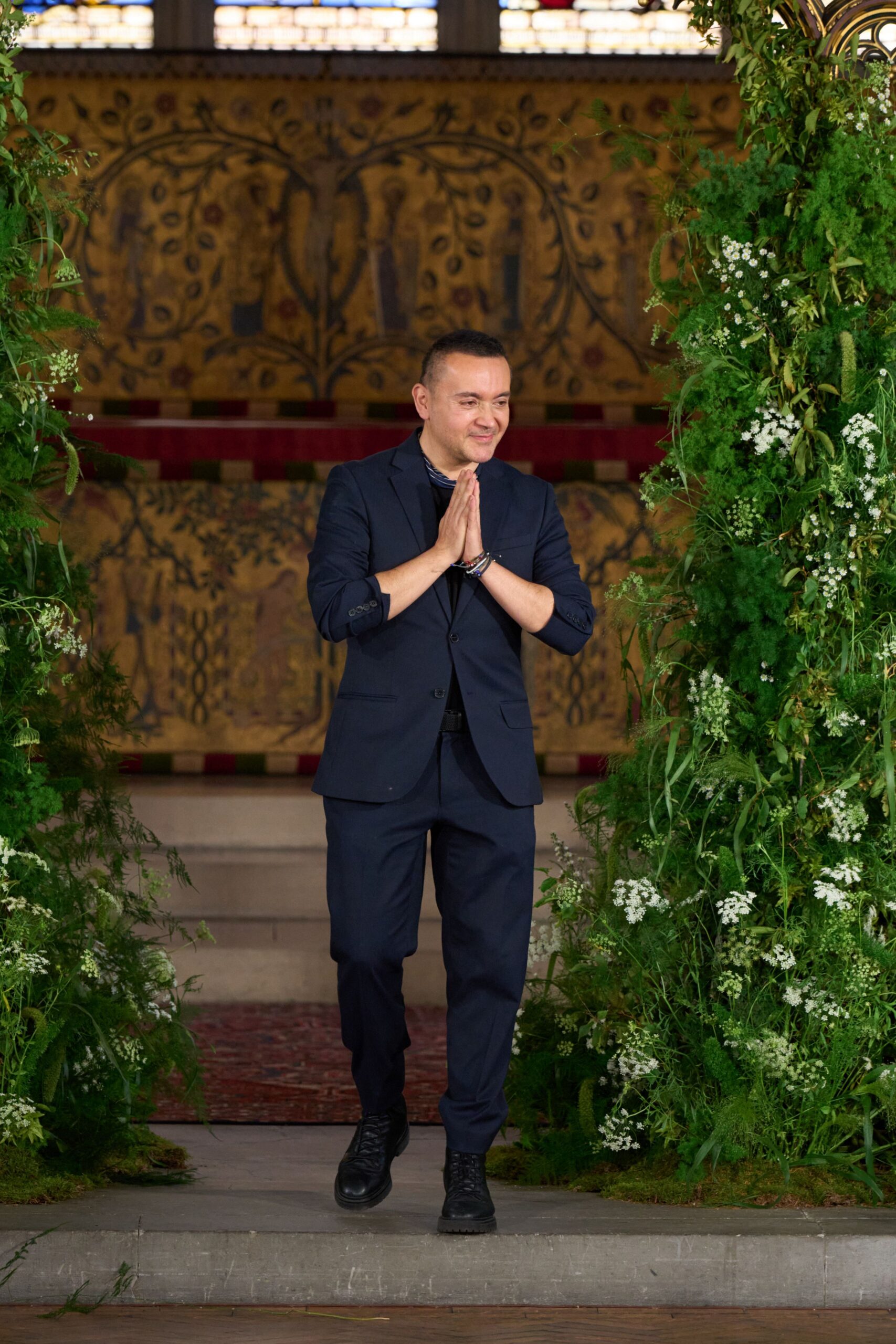 Ink-navy wool blazer with narrow lapels and a two-button front. Black crewneck tee sitting close under the jacket. Matching tailored trousers with a plain straight fall. Black leather lace-up boots with thick lug soles. Stacked cord and bead bracelets at one wrist. Stepping out beneath church greenery after the last bow.