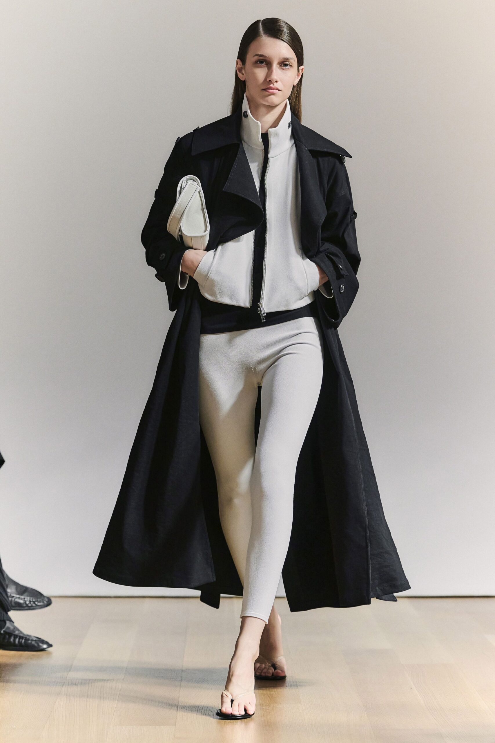 Black cotton trench, cut long with a broad storm flap. White funnel-neck jacket, silver zip ending in a tiny ring pull. Bone stretch leggings, smooth and almost matte. White folded clutch tucked under the arm. Barely there black thong sandals. For crossing a museum atrium before the rain starts again.