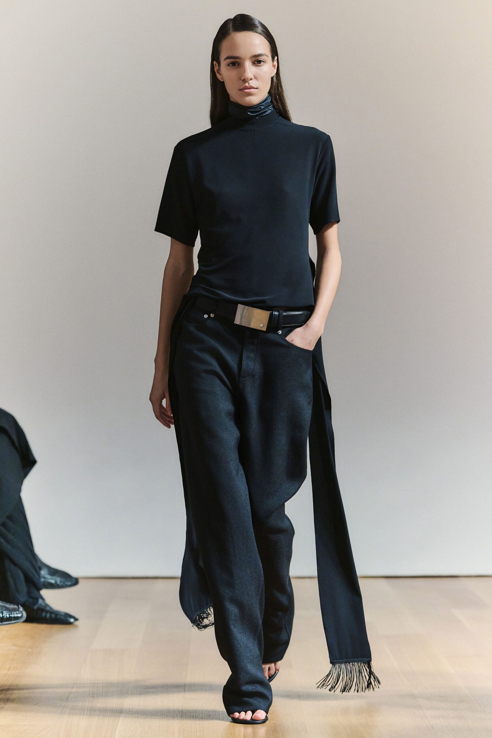 Ink-black jersey top with a softly twisted funnel neck. Black trousers cut loose through the leg, one scarf panel trailing in fringe. Wide black belt with a brushed rectangular plaque buckle. Barely there black thong sandals. For crossing a silent lobby before anyone else is downstairs.