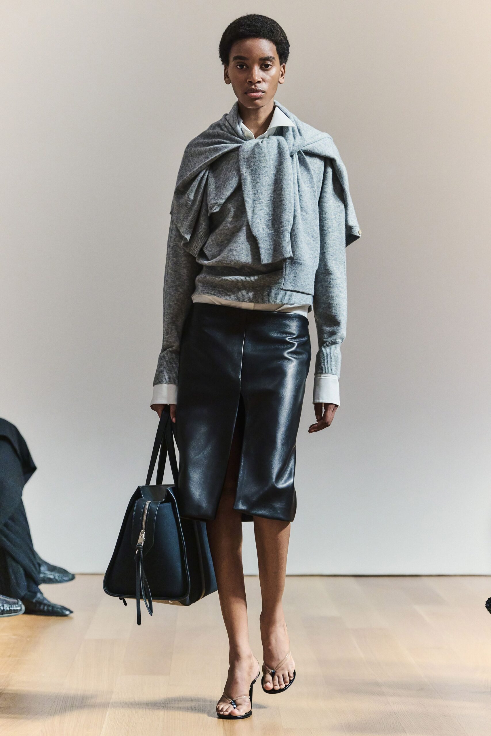 White cotton shirt, collar sharp above the knit. Heather-grey crewneck with a second sweater knotted across the shoulders. Black leather pencil skirt split high at the front. Structured black tote with a long zip tassel. Black thong kitten heels. For the first meeting after lunch, carrying too much and refusing the coat check.