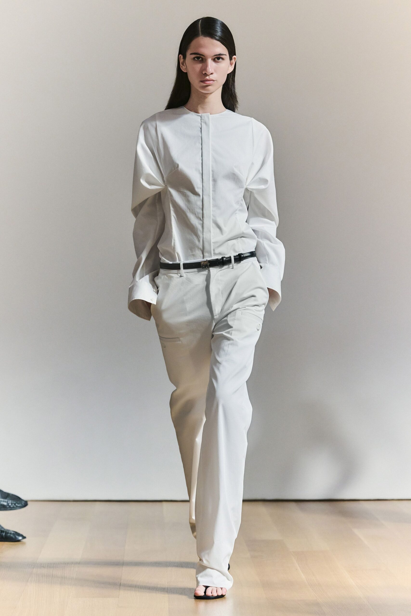 White cotton blouse with hidden placket and sculpted sleeves collapsing at the cuff. Thin black leather belt with a small square metal clasp. Ivory tailored trousers, one flap pocket set low on the thigh. Black thong sandals peeking under the pooling hem. For crossing a museum atrium before anyone else has arrived.