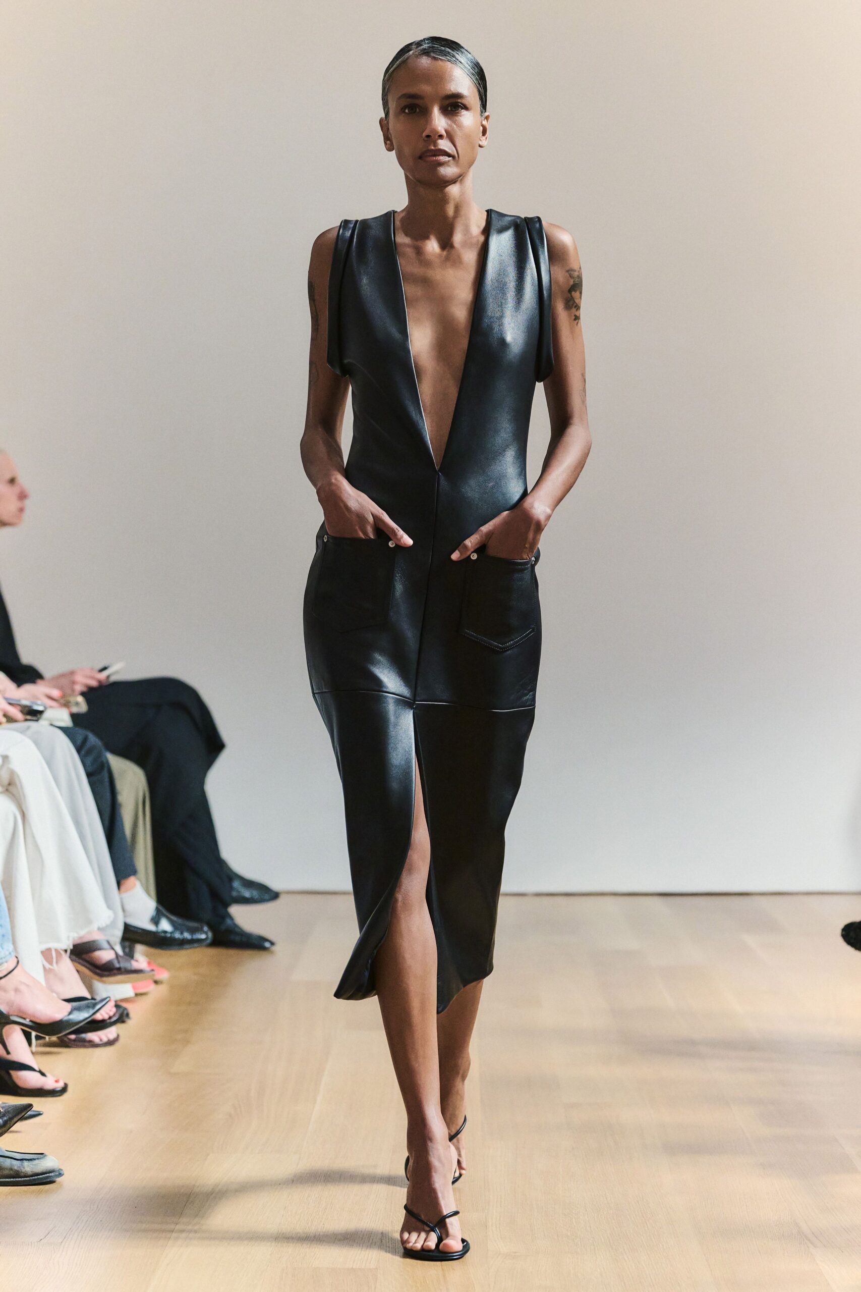 Black leather sheath dress with a plunging V and riveted patch pockets. Barely-there black thong straps on a flat square sole. For crossing a sunlit lobby after signing papers you may regret.
