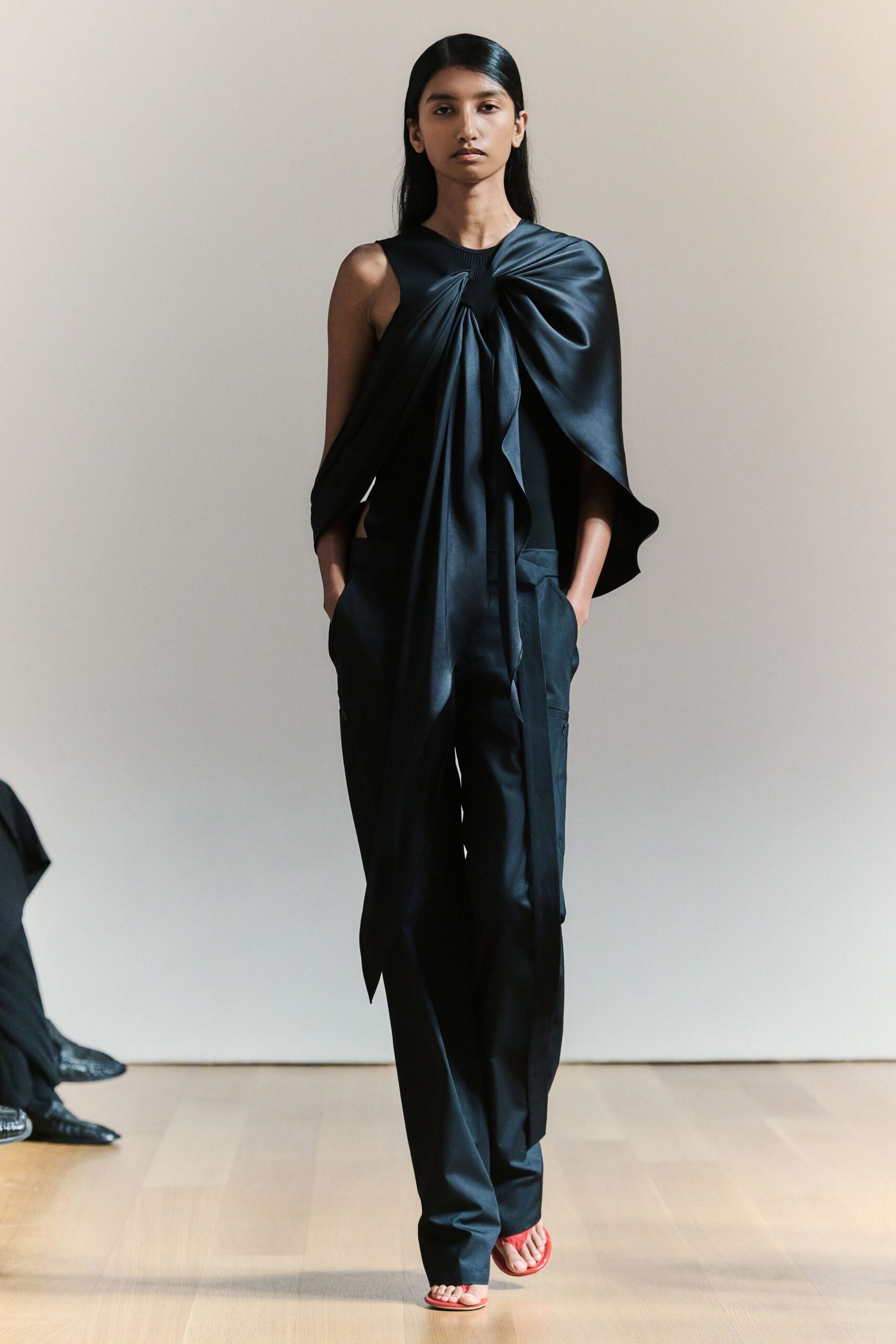 Black satin top knotted at one shoulder, one side falling cape-like. Black straight trousers with flap cargo pockets and a long hanging panel. Flat red thong sandals. Crossing a museum atrium after rain, carrying your shoes in your hand.
