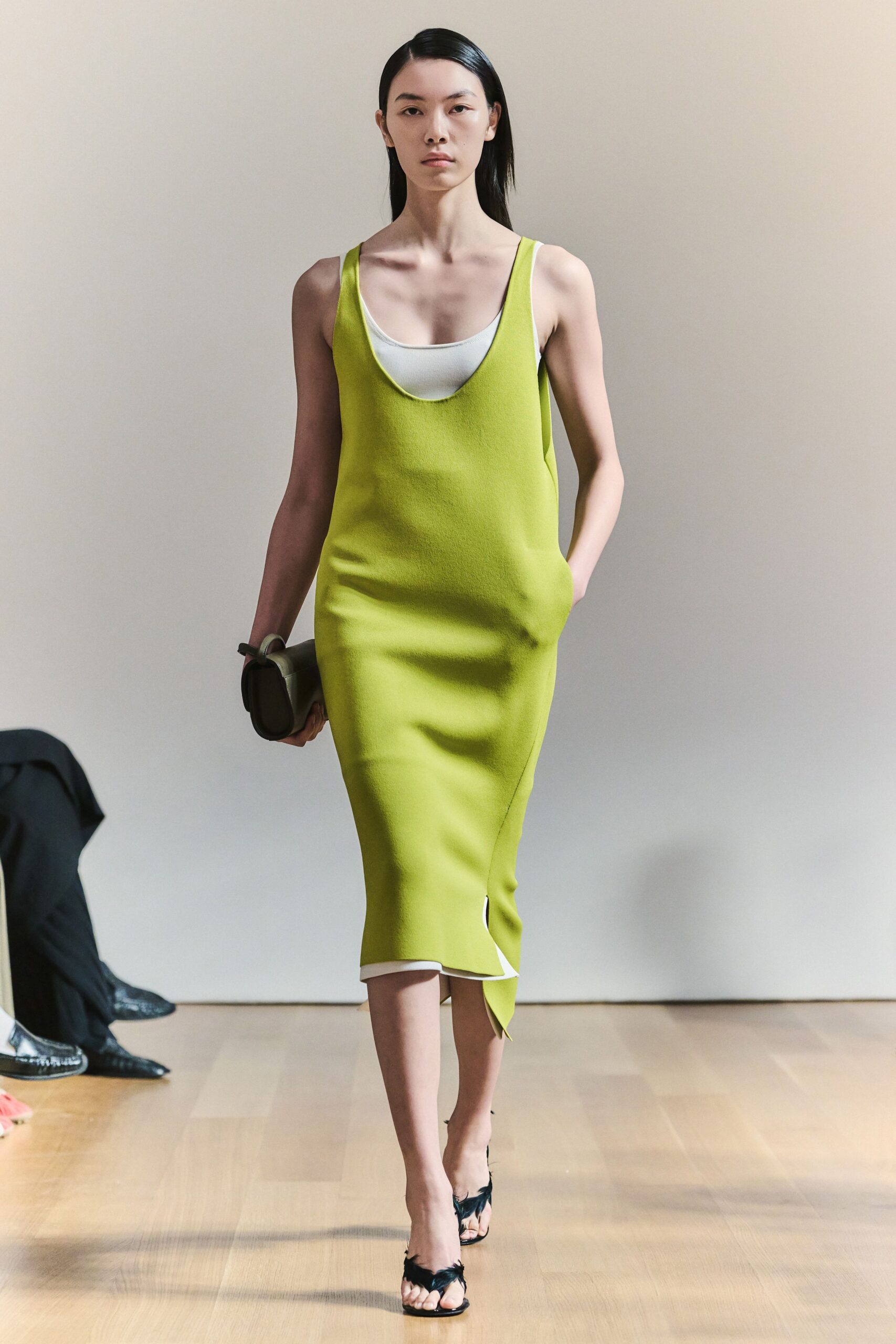 Chartreuse crepe slip dress, scooped low over a white inset layer. Olive box clutch with a looped strap at one end. Black thong sandals trimmed with wispy feathers across the toes. Gallery lunch in a white room, sunlight sliding across the floorboards.