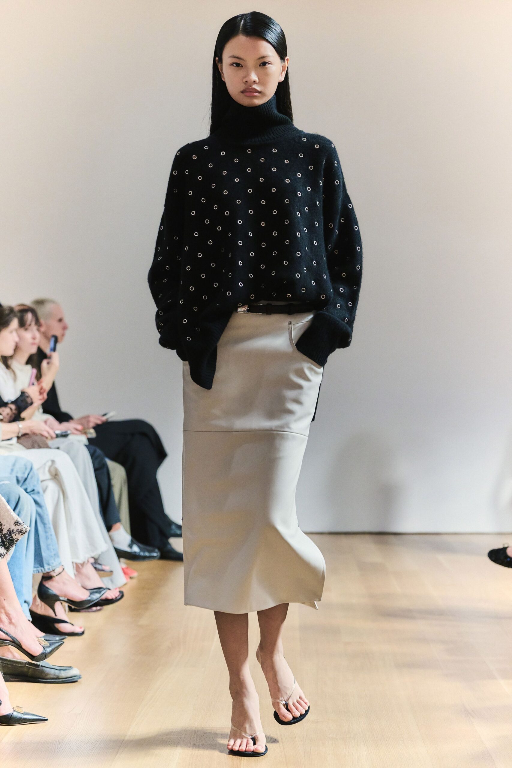 Black knit turtleneck scattered with tiny silver eyelets, one cuff swallowing the hand. Narrow black belt with a small silver clasp. Putty leather midi skirt, hip zip curving off-center above a dropped seam. Clear thong sandals with black soles. For the first cold day when the office windows finally stay shut.