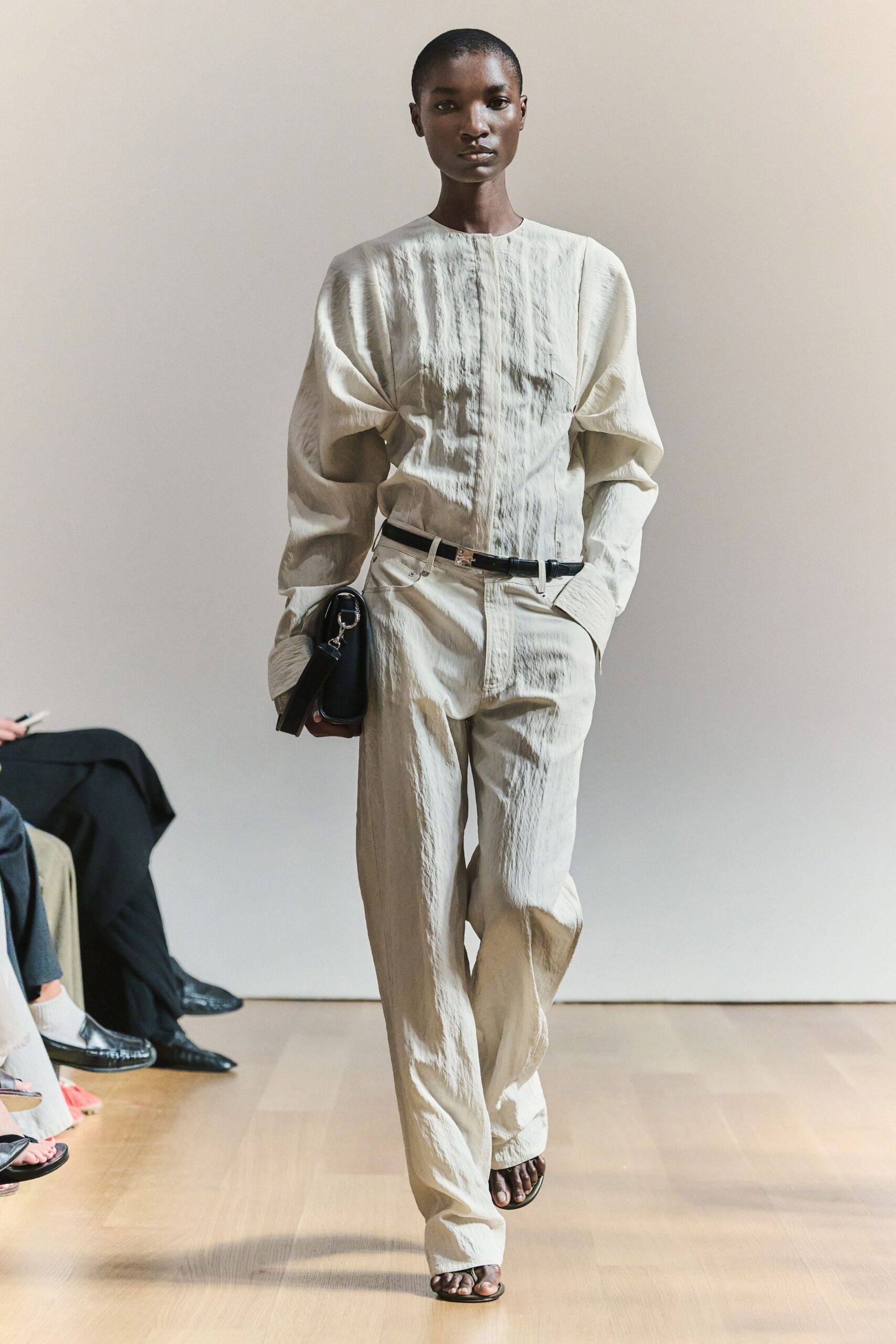 Bone crinkled blouse with cutout elbows and a concealed placket. Matching straight trousers in the same paper-crisp cloth. Narrow black belt with a small silver clasp. Black boxy shoulder bag, oversized ring hardware at the strap. Black thong sandals. For crossing a hushed museum lobby just before it opens.