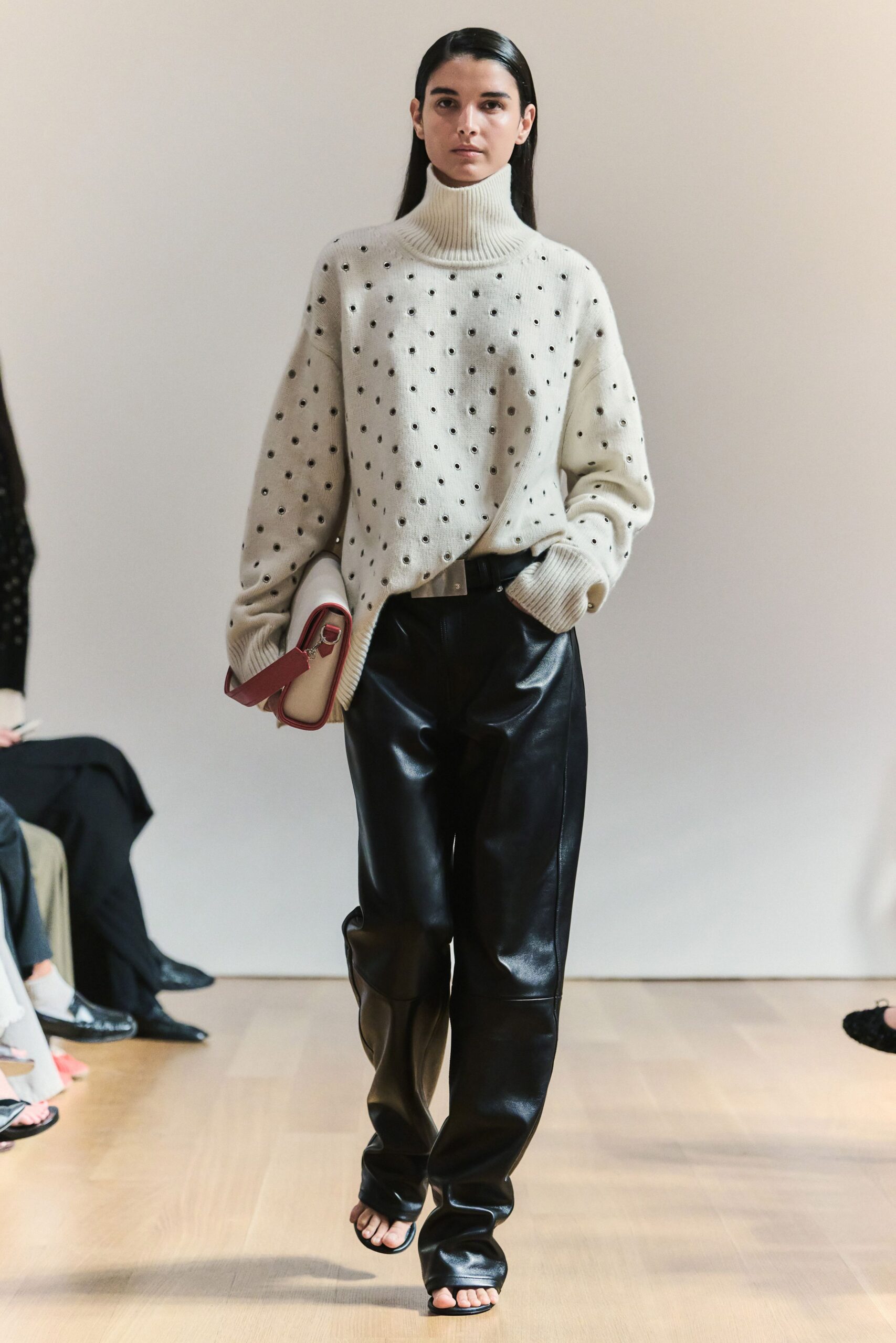 Cream ribbed knit turtleneck scattered with small metal eyelets. Black leather trousers, cut loose and pooling over the foot. Cream clutch with a wide oxblood strap and silver ring clip. Barely there black thong sandals. Gallery benefit at noon, carrying your flats home in the bag.
