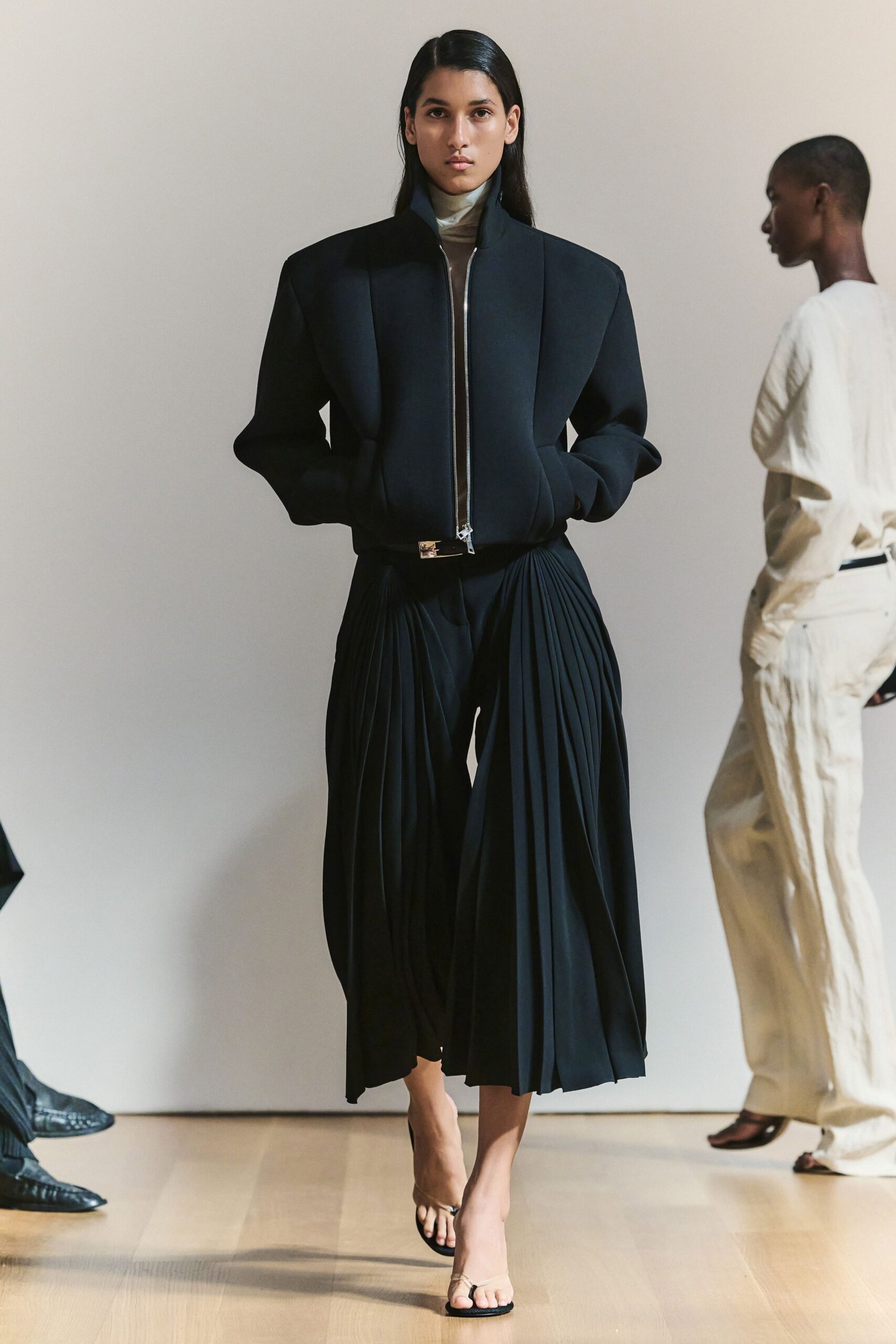 Ink black bomber with carved shoulders and a long silver zip. Sheer taupe mesh turtleneck, doubled pale at the throat. Black culottes with deep knife pleats opening from the hip. Slim black belt, bright metal clasp at center. Barely there black thong heels. Walking into a museum lobby before the rain starts again.