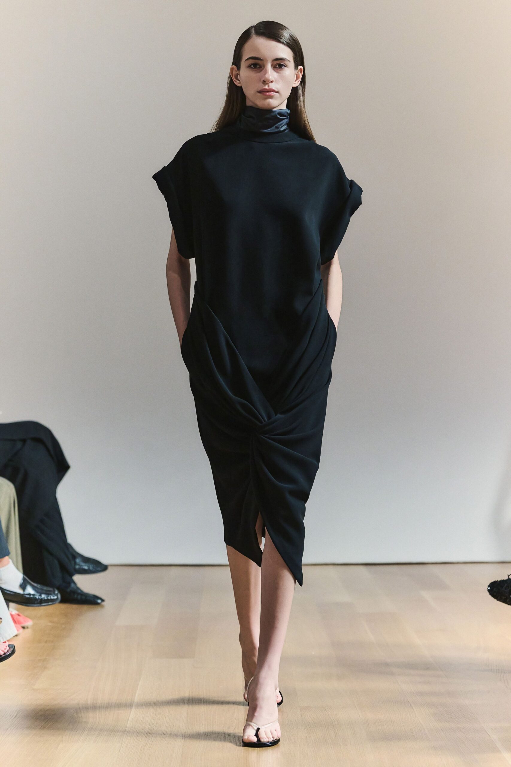Black crepe dress with a satin funnel neck and twisted drape. Bare thong sandals with a glossy black sole. Crossing a silent hotel lobby before anyone has set out breakfast.