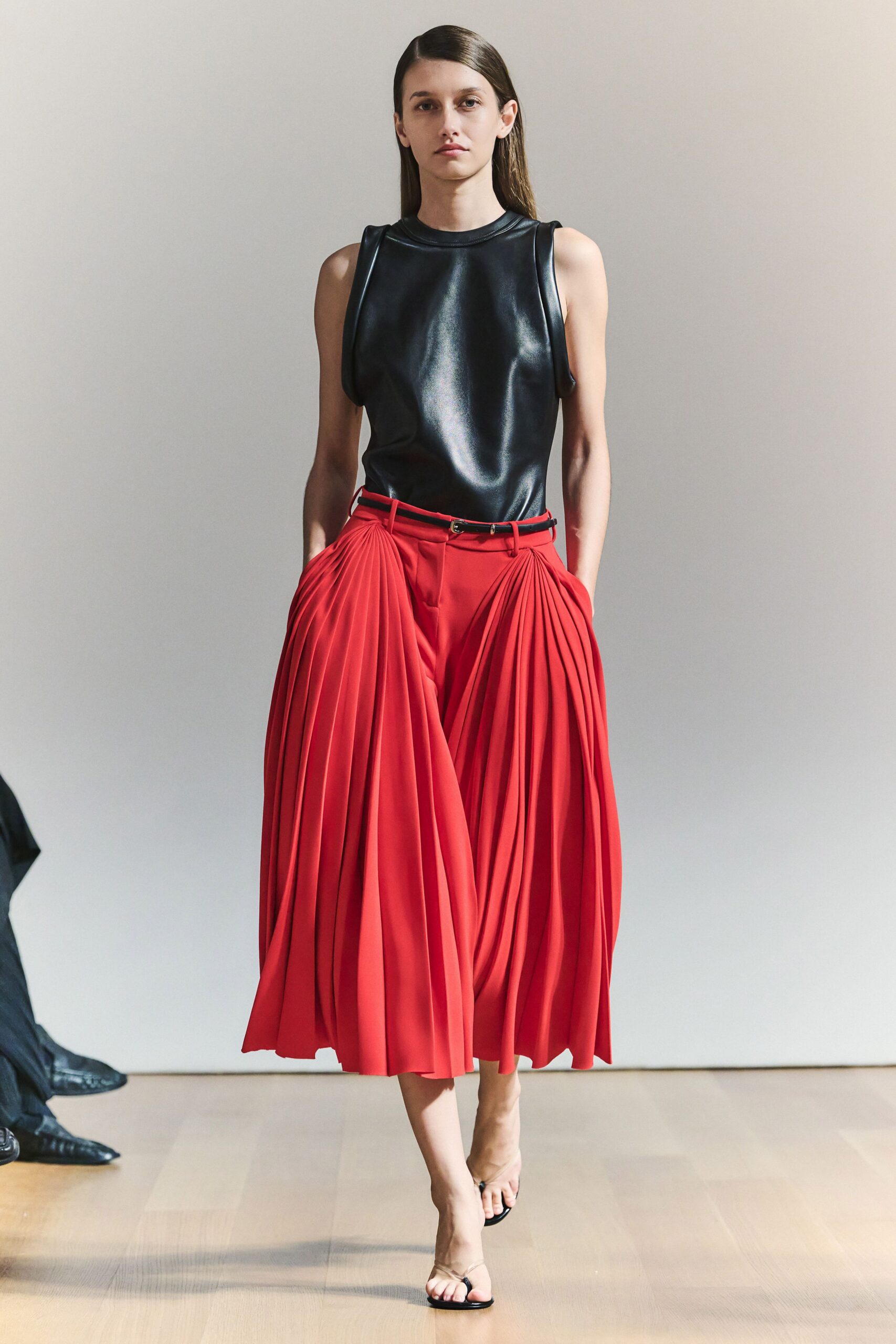 Black leather shell top with rolled armholes and a high crew neck. Thin black belt, tiny oval buckle sitting slightly off center. Tomato red culottes with deep pleated panels swelling from the hips. Barely there black thong sandals. Crossing a museum atrium before the air-conditioning has fully kicked in.