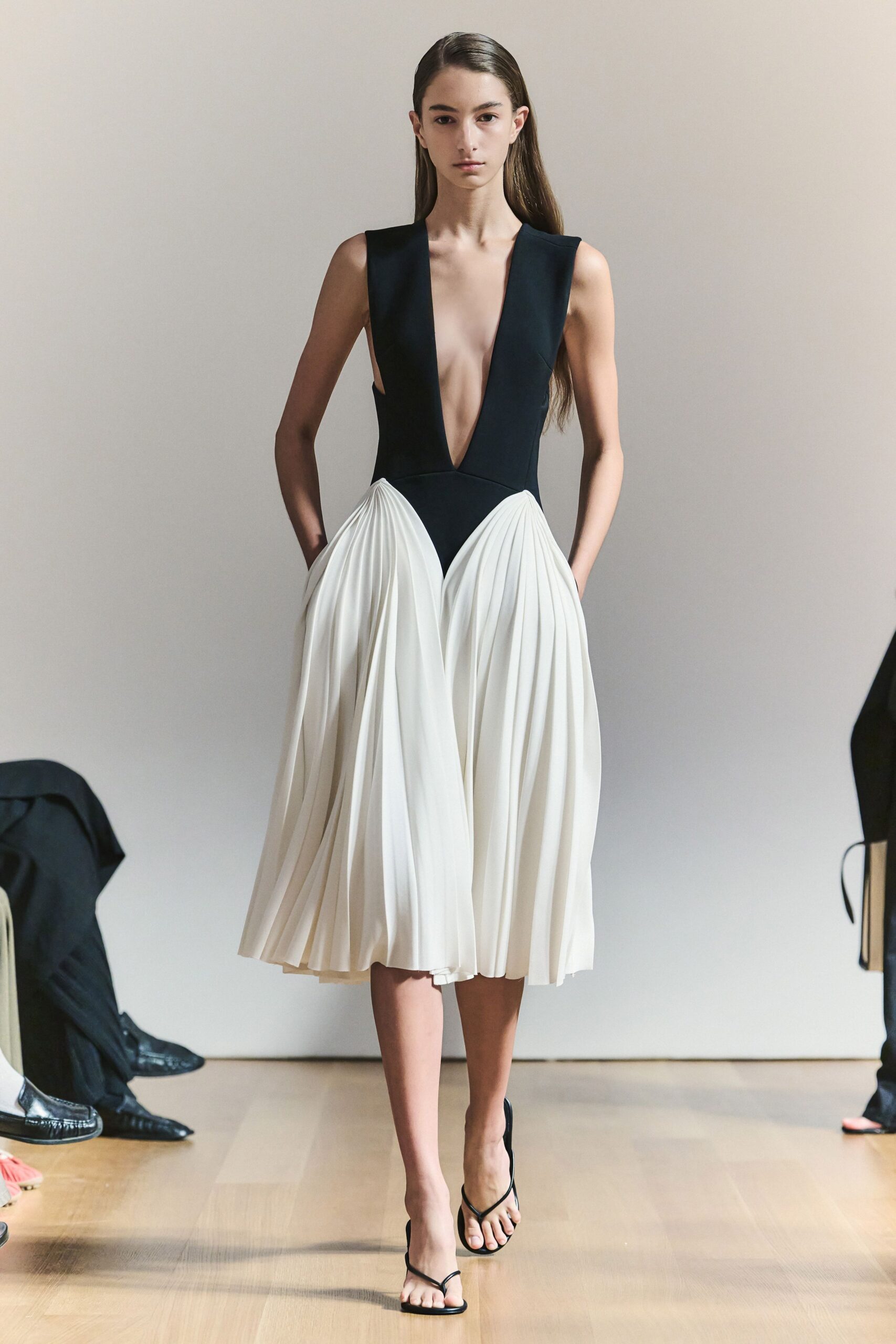 Black bodice plunging to the waist, opening over a white knife-pleated skirt. Bare black thong sandals with a slim slingback strap. Walking across a rented apartment's wood floor before the florist arrives.