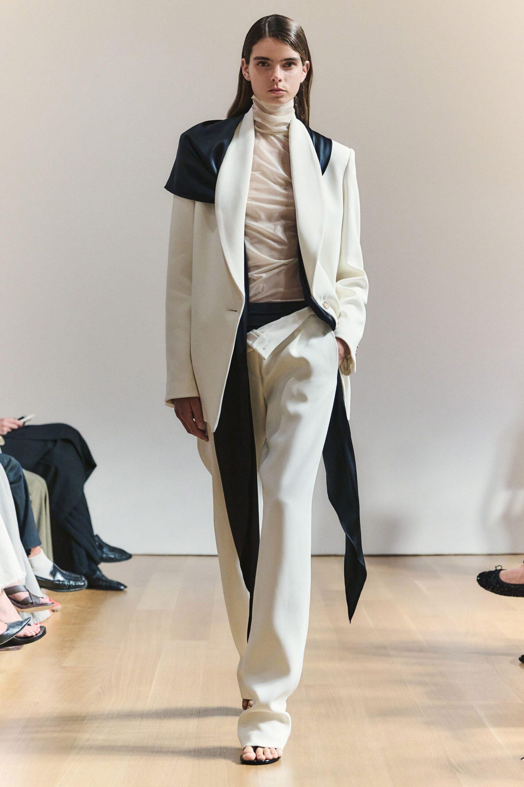 Sheer oyster jersey turtleneck, softly ruched through the torso. Ivory tailored blazer with black satin shoulder panels and lapel lining. Ivory wide-leg trousers, wrapped panel jutting sharply from the waist. Black thong sandals barely visible under the pooled hem. For crossing a museum atrium before the donor lunch begins.