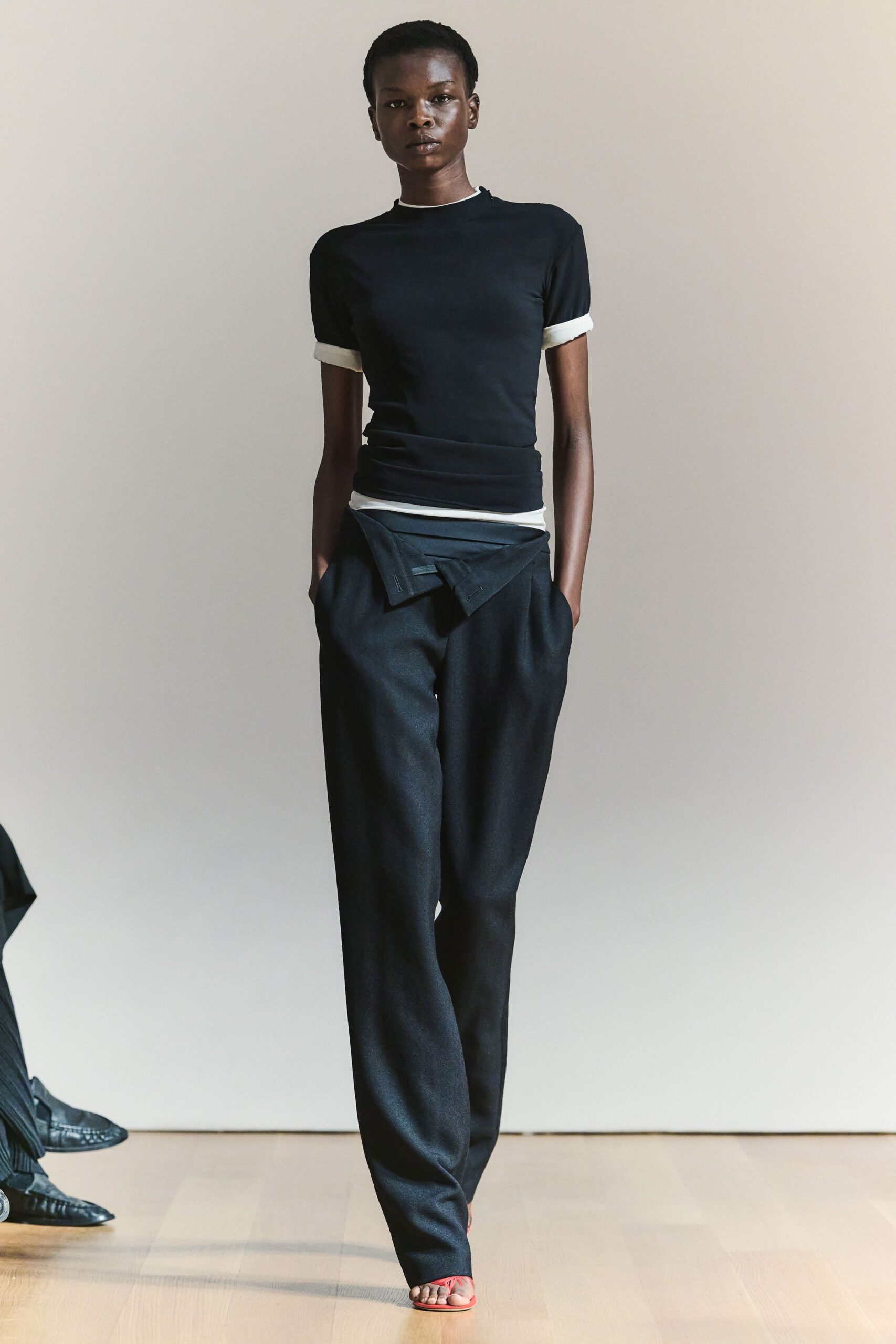 Black stretch tee with white binding at the neck and rolled cuffs. White jersey band peeking out at the waist. Black wool trousers cut full through the leg. Inside-out trouser panel folded into a sharp hip drape. Flat coral thong sandals. Walking into a bare studio while the paint on the skirting boards dries.