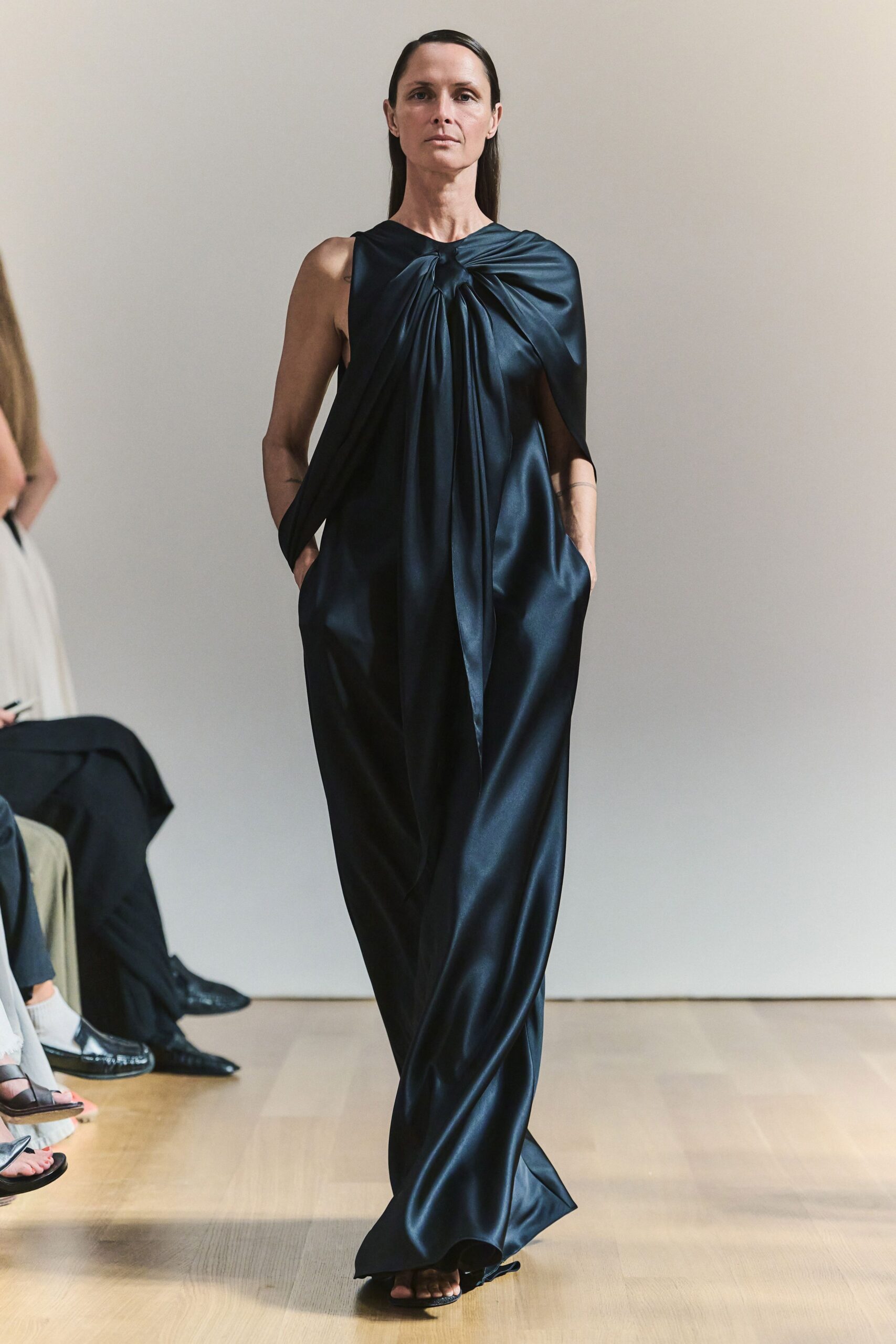 Black satin column gown, twisted at the chest into a long central drape. Barely there black sandals under the pooling hem. For crossing a museum atrium after everyone else has left.