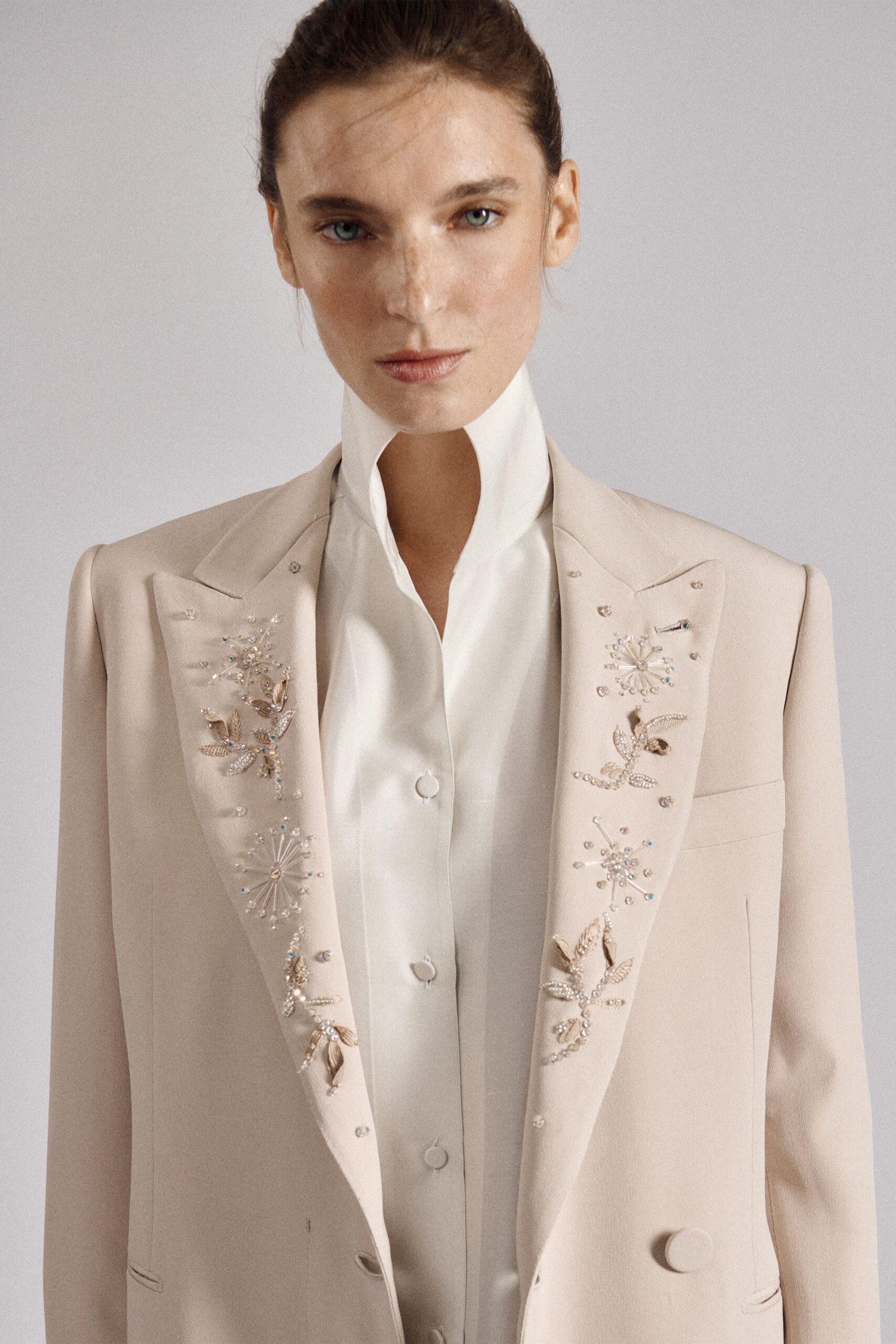 Powder blush tailored blazer, lapels scattered with crystal starbursts and metallic petals. Ivory satin shirt with a high split standing collar and covered buttons. Registry office noon, fountain spray catching on the courthouse steps.