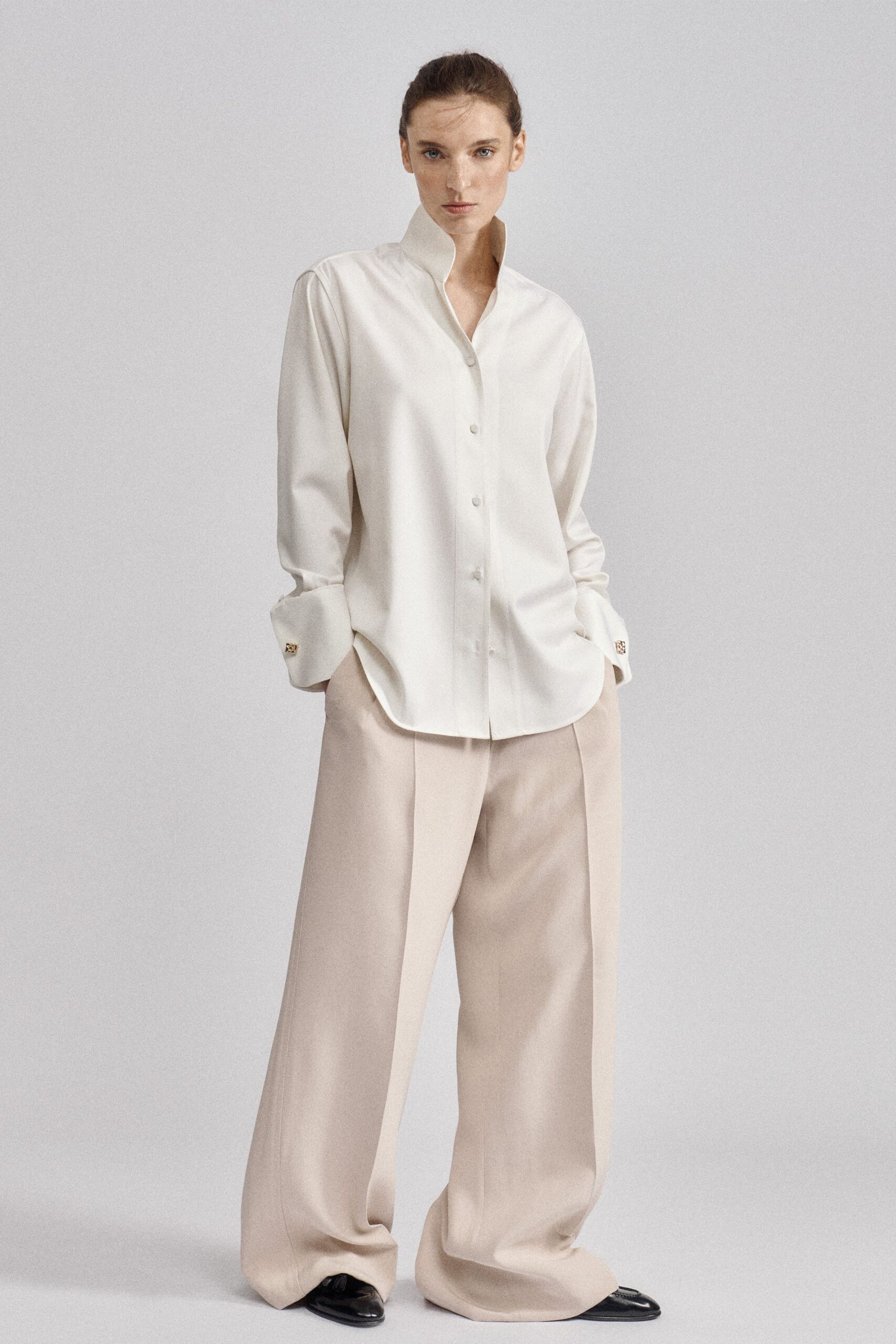 Ivory satin shirt with a high standing collar and turned-back cuffs. Pale blush wide-leg trousers, pressed front crease breaking over the shoe. Black patent loafers peeking from the pooling hem. Early ferry crossing, coffee cooling before the city comes into view.