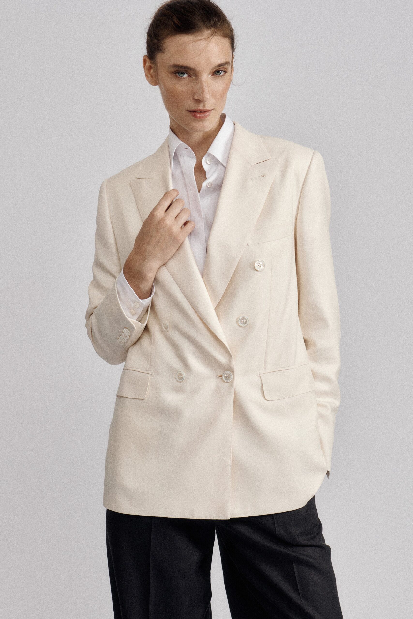 Cream double-breasted blazer with peaked lapels and glossy tonal buttons. White cotton shirt, top buttons left open beneath the jacket. Black tailored trousers with a clean flat front. Early train to a civil wedding, coffee cooling on the platform.