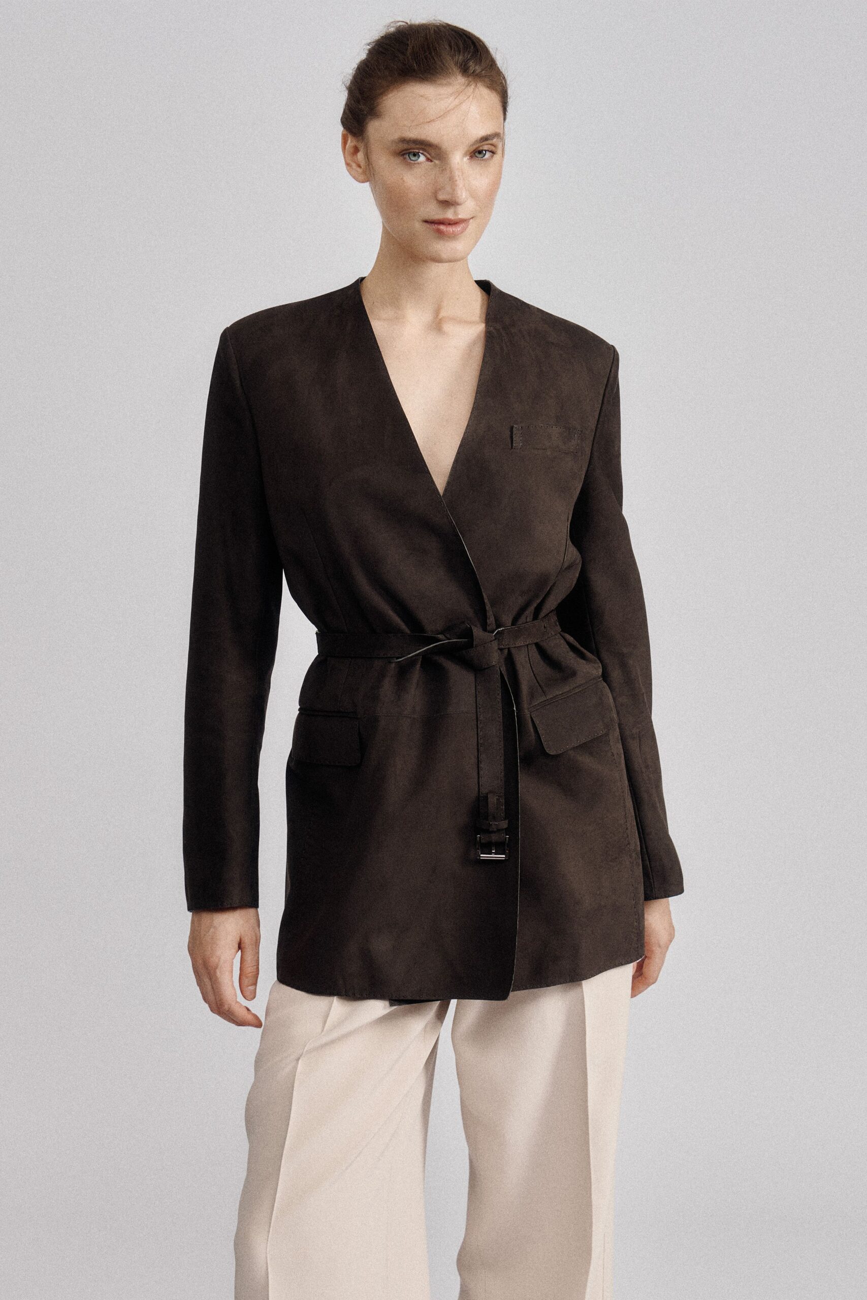 Espresso suede wrap coat, deep V front and belt buckle hanging low. Cream wide-leg trousers with a pressed crease falling clean from the hip. For a quiet lunch where the table linen matters more than the menu.