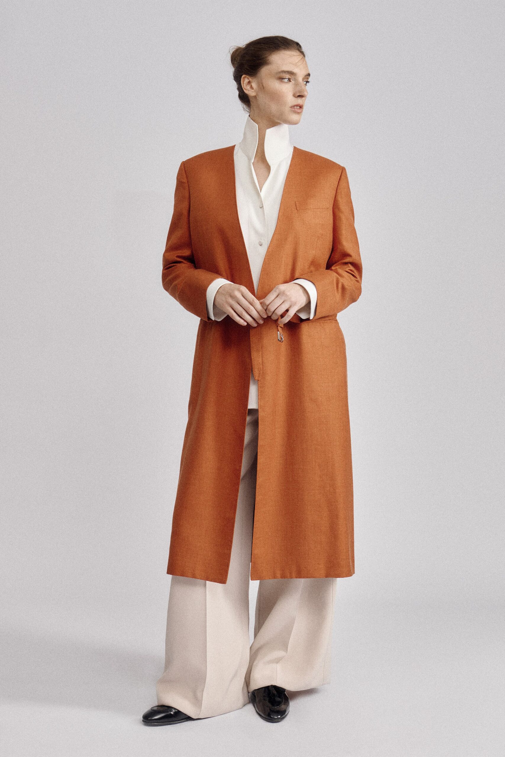 Burnt cinnamon long coat with a deep collarless plunge and dangling tab. Ivory shirt with a cutaway standing collar, buttons disappearing below the chest. Cream wide-leg trousers breaking softly over the shoes. Black leather loafers with creased, mirror-bright toes. Museum atrium at opening hour, heat still trapped in the stone.