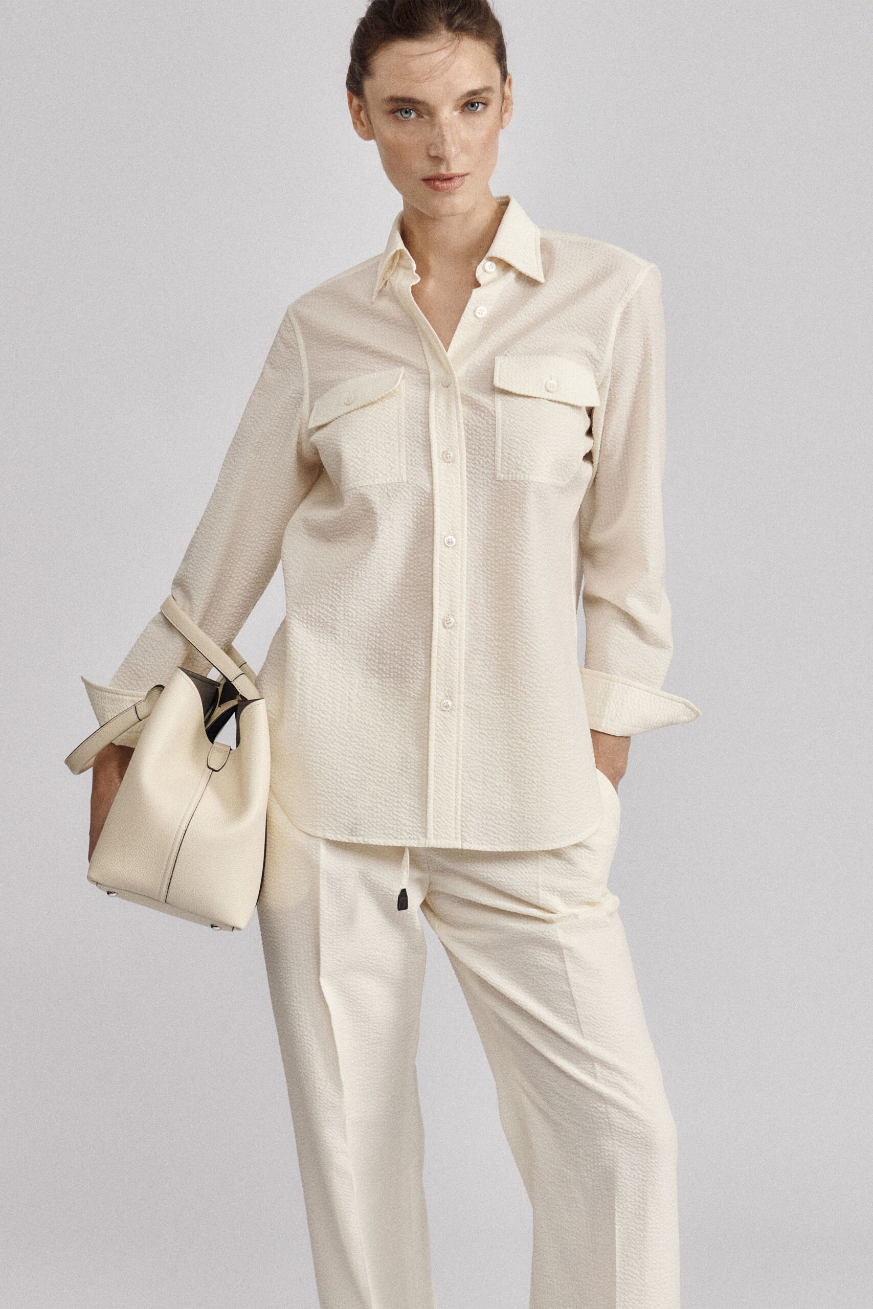 Ivory pebbled shirt with flap chest pockets and a softly rolled cuff. Matching straight trousers, drawstring tipped with a small black aglet. Cream leather bucket bag with slim straps and silver feet. For the first ferry over, when the air still smells of rope.