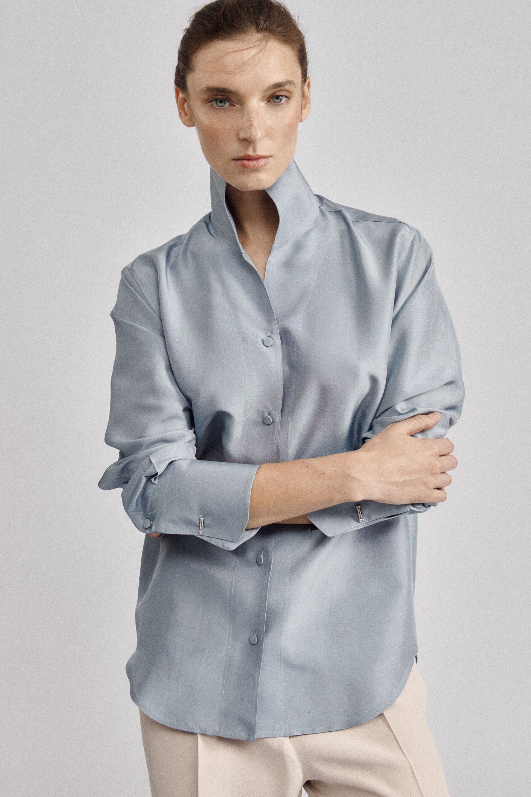 Powder-blue satin shirt, high stand collar and fabric-covered buttons. Cream tailored trousers with a soft pressed crease. Morning meeting room, coffee gone cold beside a stack of printouts.