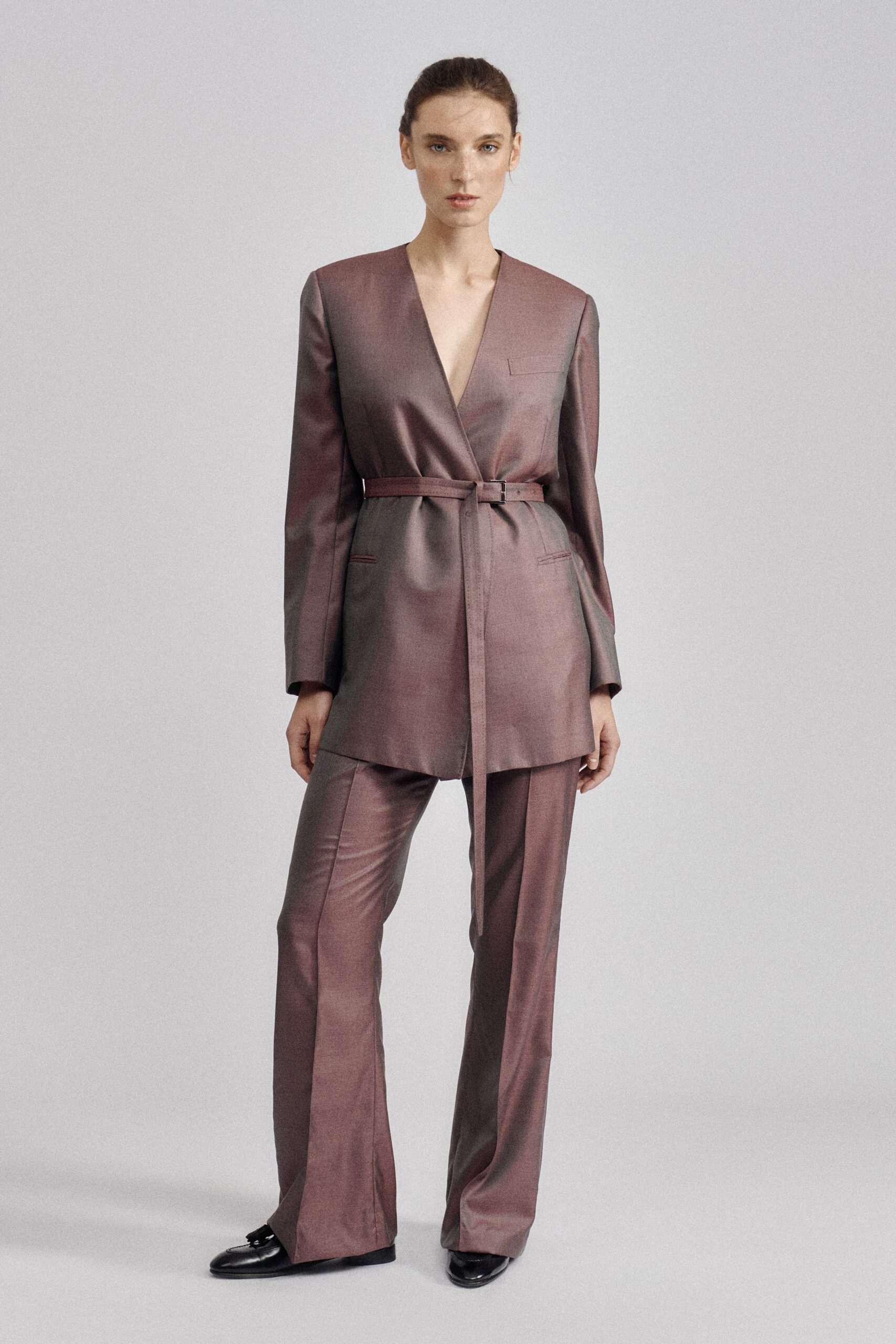 Mauve-brown satin jacket cut long, with a plunging collarless wrap front. Self-fabric belt with a small rectangular buckle and one long hanging end. Matching satin trousers, pressed creases breaking softly over the shoes. Black loafers with a tasseled vamp. For the meeting after dusk when the office windows turn mirror-dark.