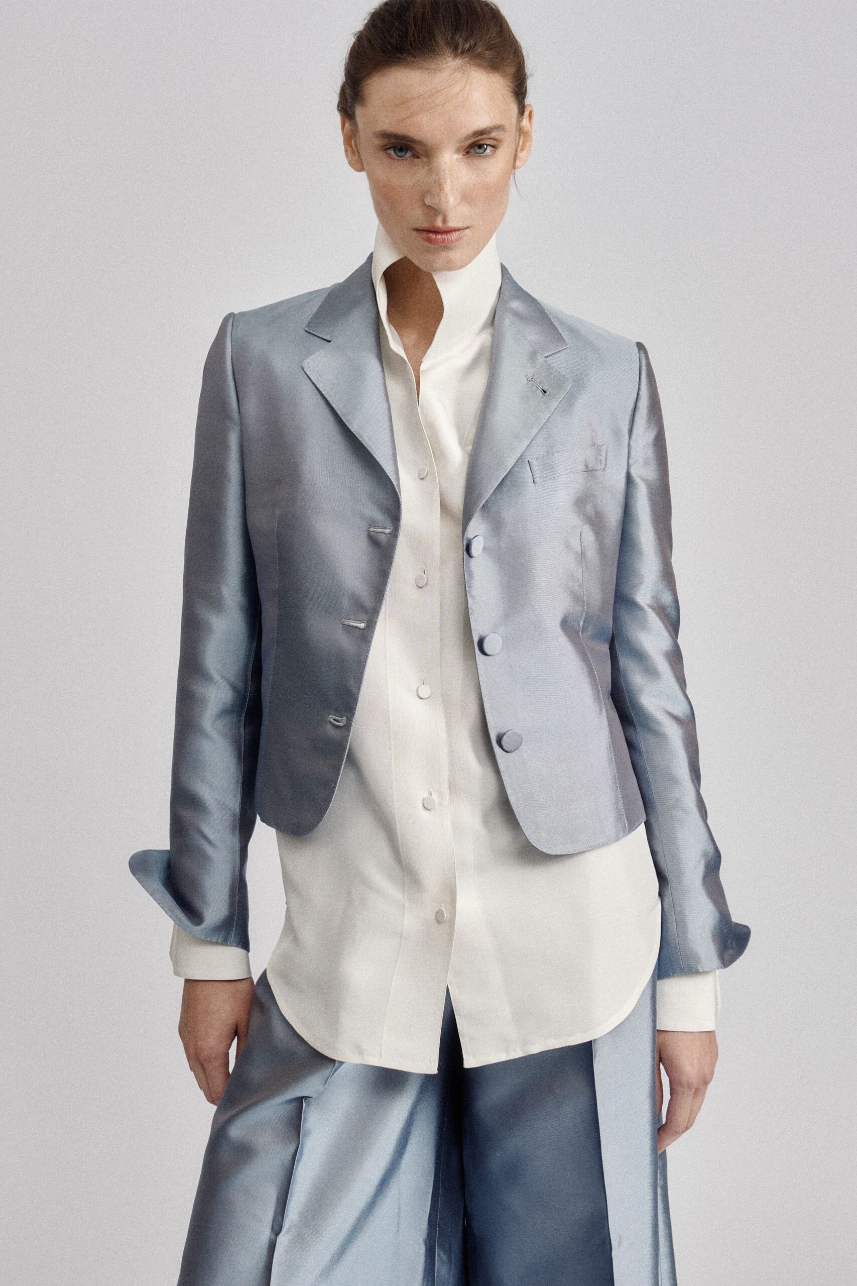 Powder-blue satin blazer, covered buttons and a tiny hand-tacked lapel stitch. Ivory shirt with a towering split stand collar and covered placket. Matching satin wide-legs, breaking into a soft puddle at the hip. Hotel corridor at dawn, carrying your shoes and yesterday's keycard.
