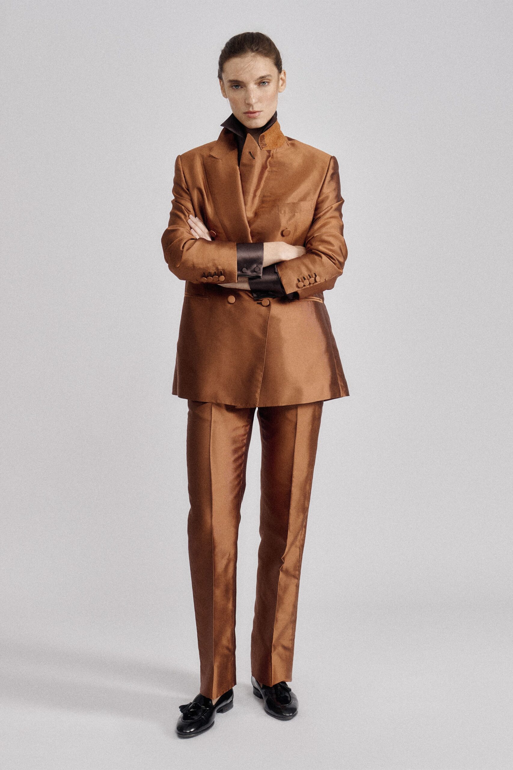 Black jersey turtleneck, snug and stark at the throat. Copper satin double-breasted blazer, one collar point kicked sharply upward. Matching slim trousers with a hard crease catching the light. Black tassel loafers, softly wrinkled over the vamp. For the hour before a committee vote in a paneled municipal office.