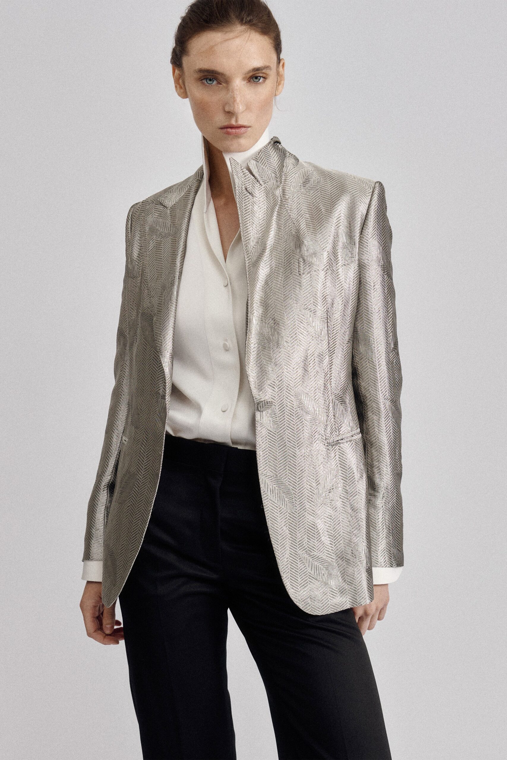 Silver herringbone blazer woven with raised leaf motifs, one lapel flipped upright. Ivory silk blouse with covered buttons and a narrow standing collar. Black satin trousers, flat-front and sharply cut through the hip. For reading the seating chart in a museum foyer before dinner.