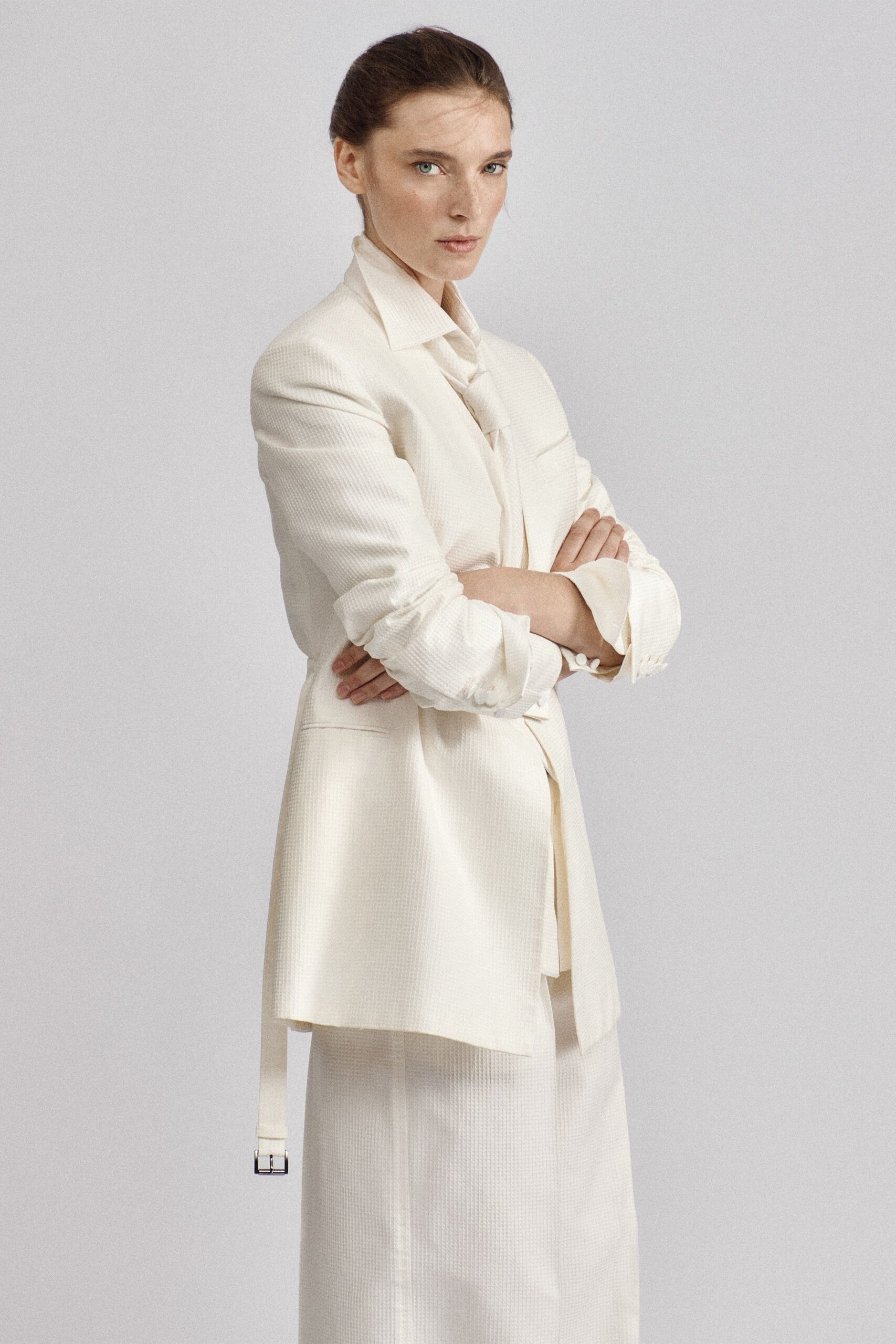 Cream micro-waffle blazer with a long belt strap hanging past the hem. Soft ivory blouse tied in a loose knot at the throat. Matching column skirt in the same faintly gridded weave. Early registry-office appointment, flowers still wrapped in brown paper.