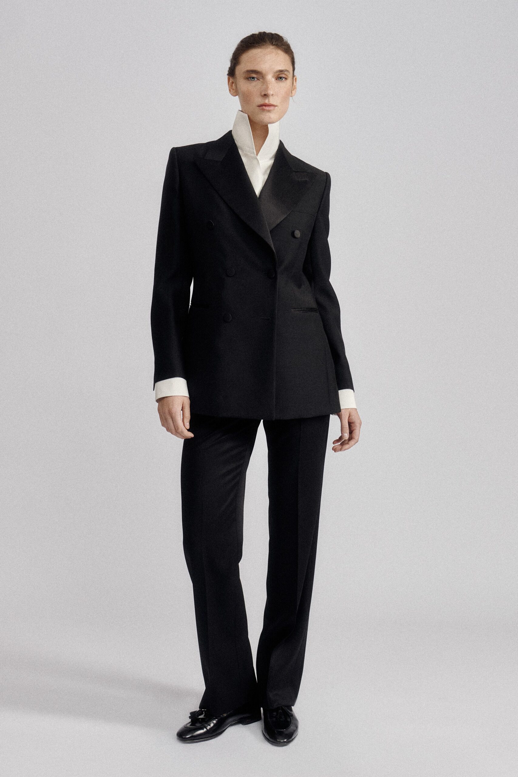 White bib-front shirt with a high standing collar framing the jaw. Black double-breasted blazer, peaked lapels and cloth-covered buttons. Black straight trousers with a pressed crease and slight break. Glossy black loafers with tasseled vamps. For reading the will in a room with the curtains still drawn.