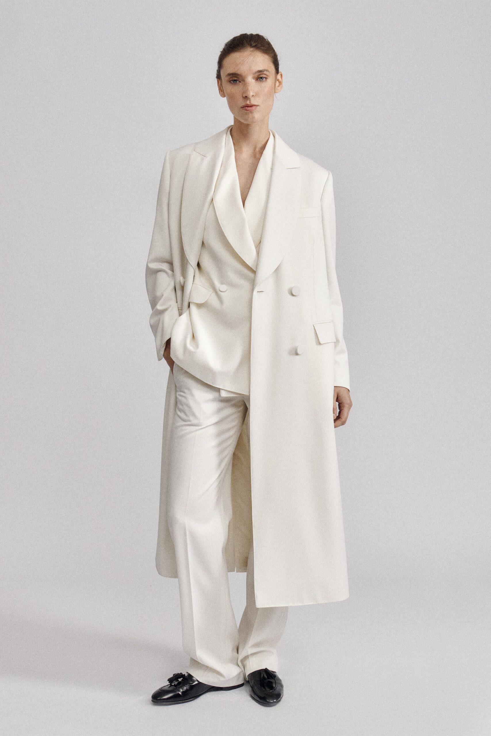 Ivory double-breasted coat, calf length with covered buttons and broad lapels. Cream satin blouse falling in a deep shawl-like drape. Ivory straight trousers with a soft break over the instep. Black tassel loafers, slightly collapsed at the vamp. For signing papers in a silent townhouse before the movers arrive.
