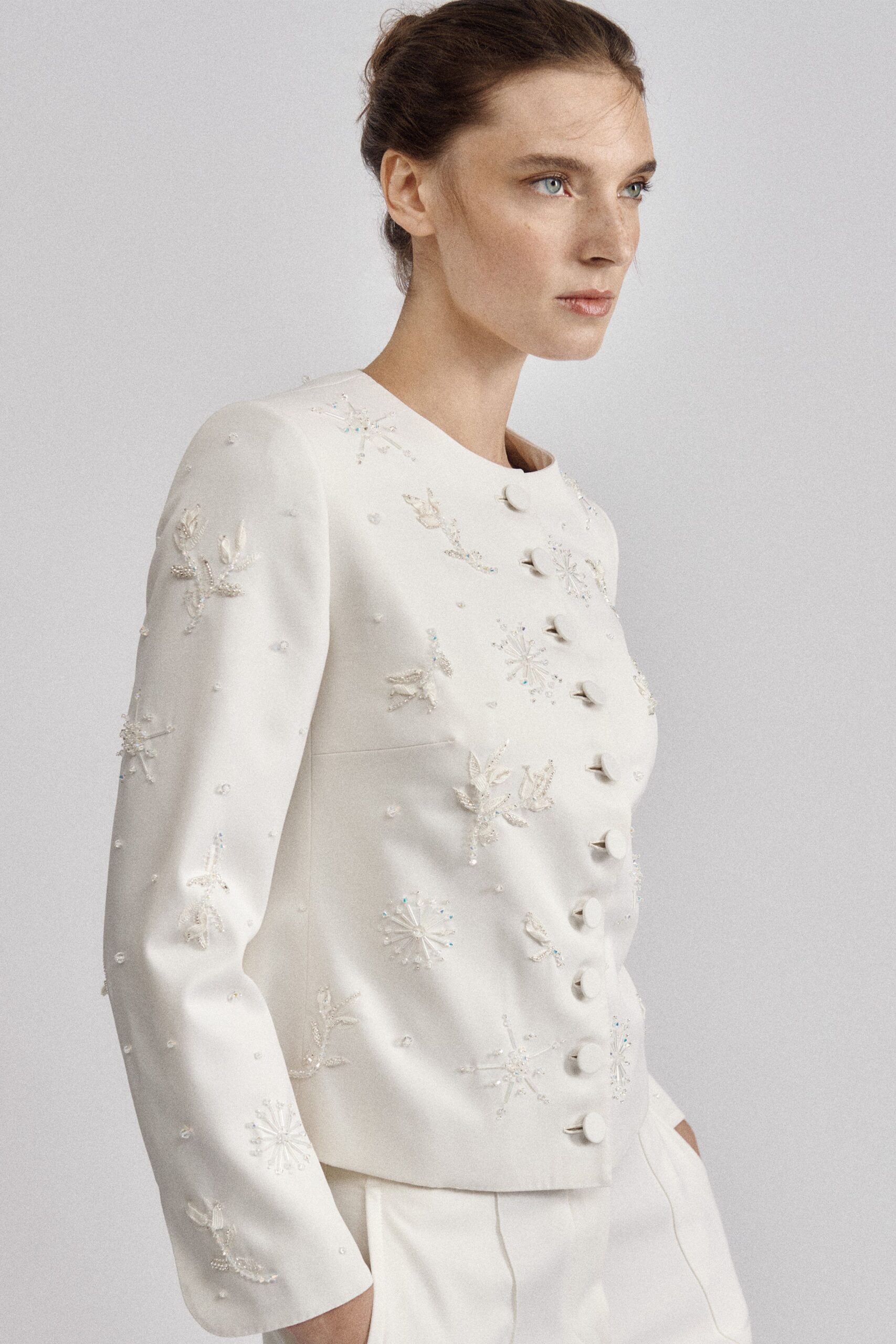 Ivory tailored jacket with padded shoulders, pearl-beaded snowflake sprays and cloth-covered buttons. Ivory straight trousers with a sharp pressed crease. City hall at noon, the bouquet still in water by the sink.
