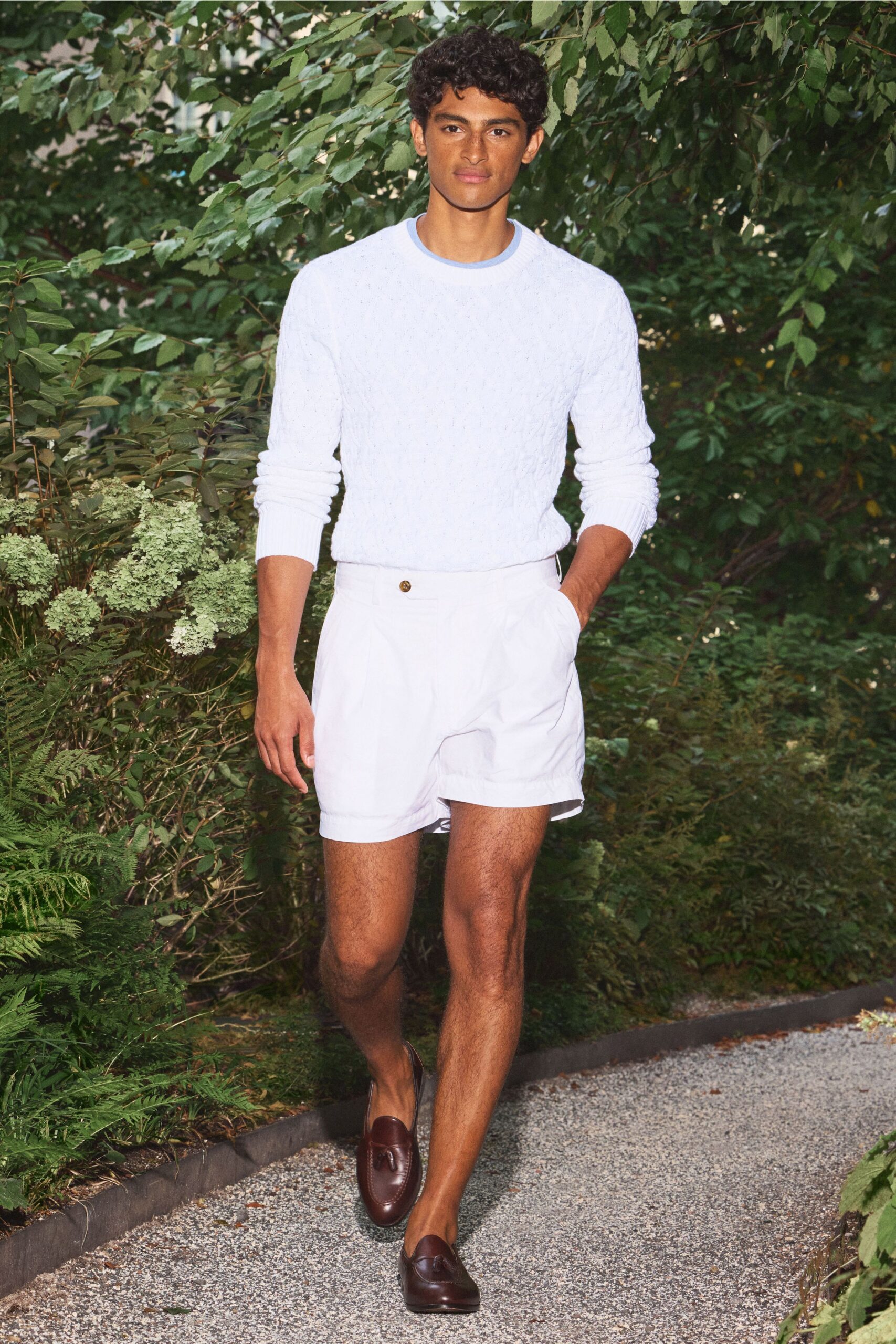 Pale blue cable-knit sweater with a neat ringer crewneck. White tailored shorts with a single horn button at the waistband. Mahogany leather tassel loafers, worn bare at the ankle. Garden path after brunch, gravel catching in the heat.