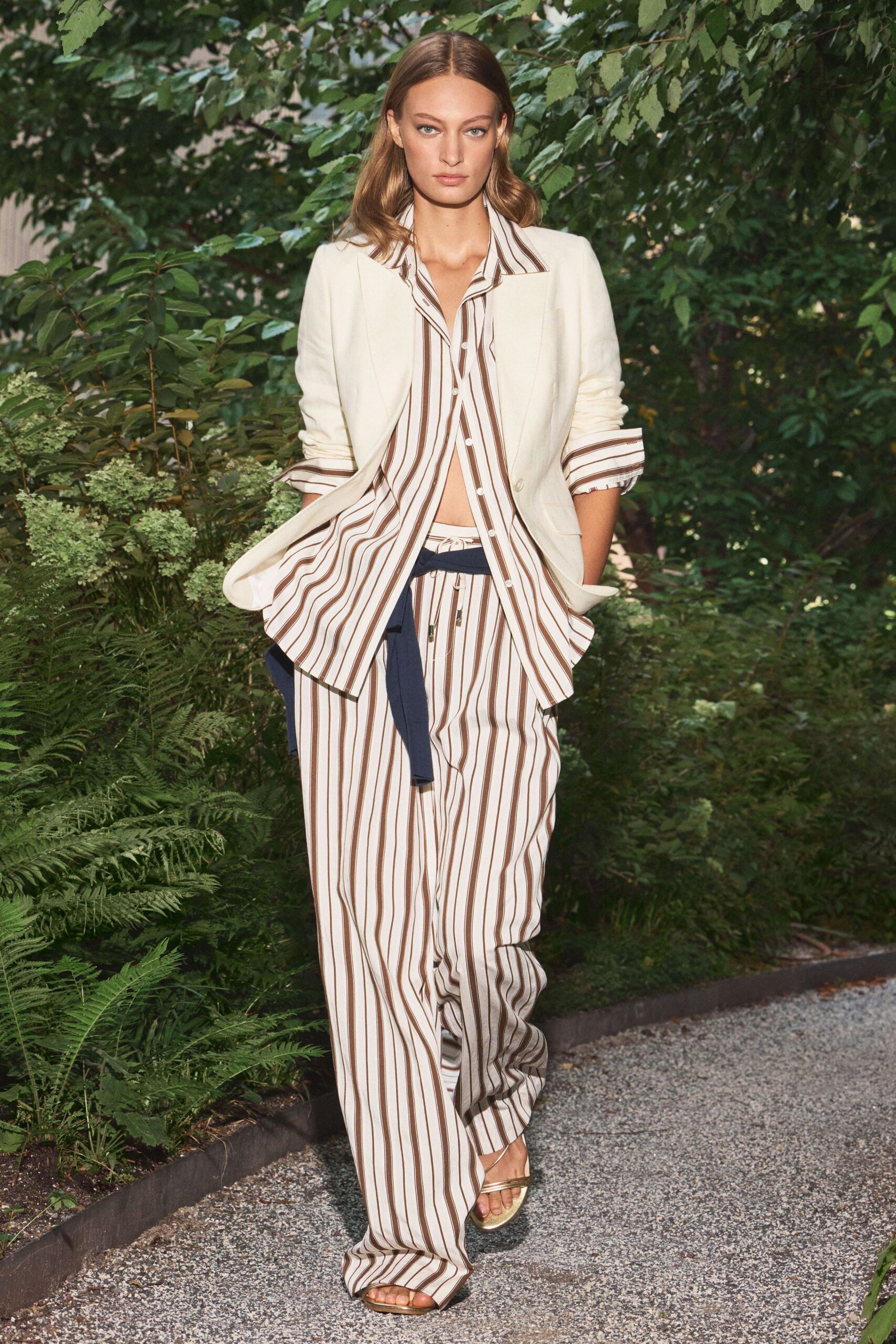 Cream blazer in a soft slub weave, sleeves pushed to the forearm. Ivory shirt with brown deck stripes, hem flaring open below the last button. Matching striped drawstring trousers, navy sash knotted low across the hips. Pale metallic slide sandals. Walking the gravel path after lunch, still carrying the hotel room key.
