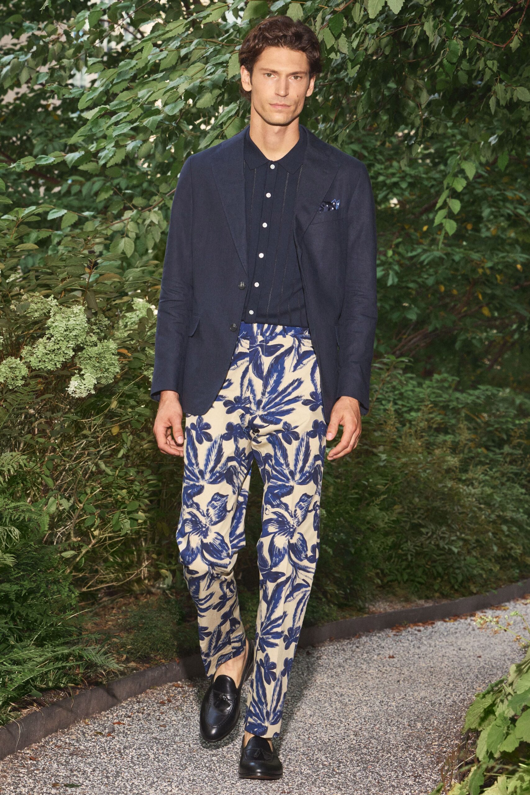 Inky linen blazer with soft shoulders and two dark horn buttons. Navy knit polo, white buttons marching between narrow stitched pleats. Cream trousers washed in oversized cobalt hibiscus brushstrokes. Blue silk peeking from the chest welt. Black tassel loafers. Walking the gravel path to a garden wedding after the heat breaks.