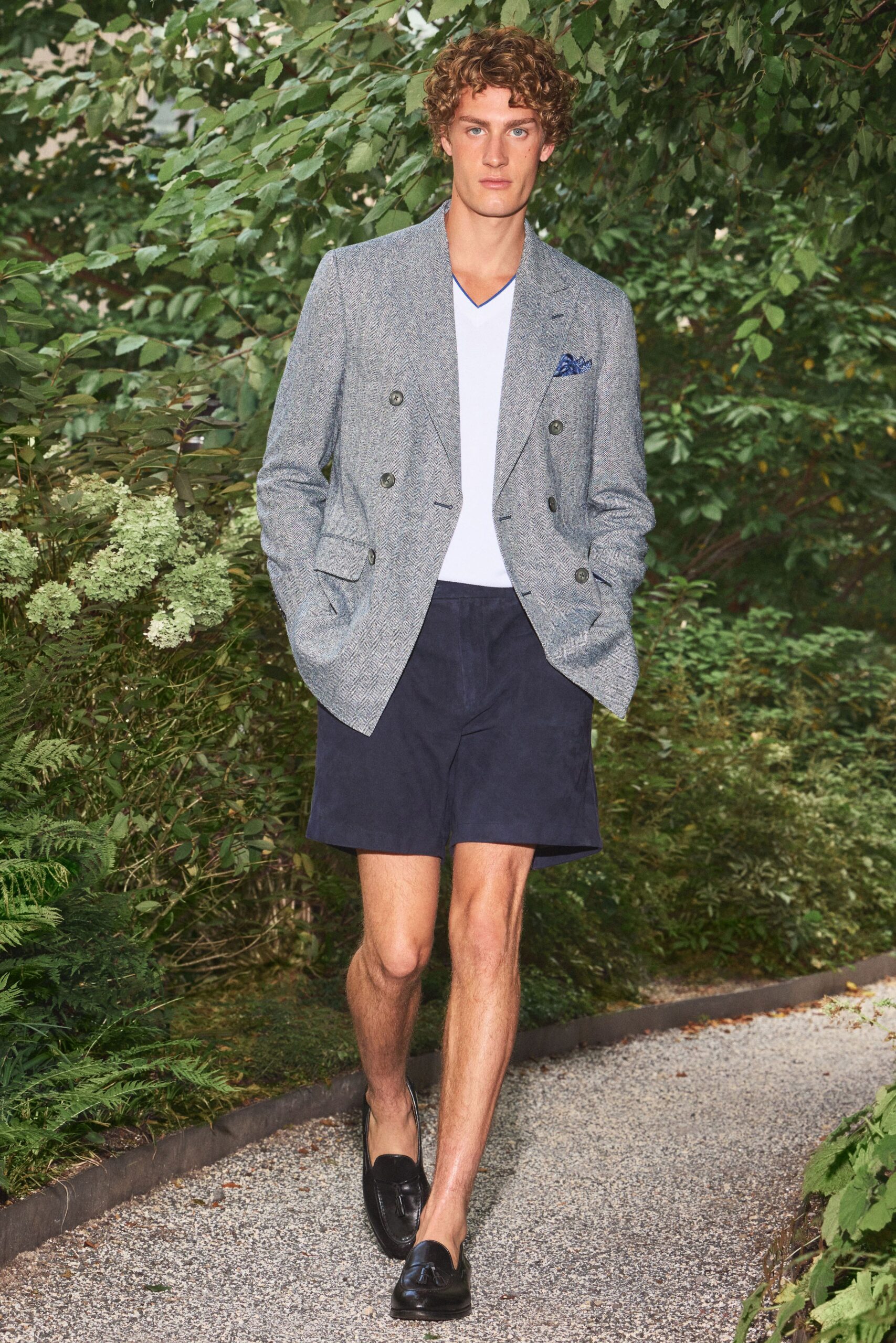 Blue-grey herringbone double-breasted blazer with dark horn buttons and a pocket square. Pale V-neck tee edged with narrow cobalt binding. Ink shorts cut full through the leg. Black leather tassel loafers. Walking the gravel path to lunch at a country house after rain.