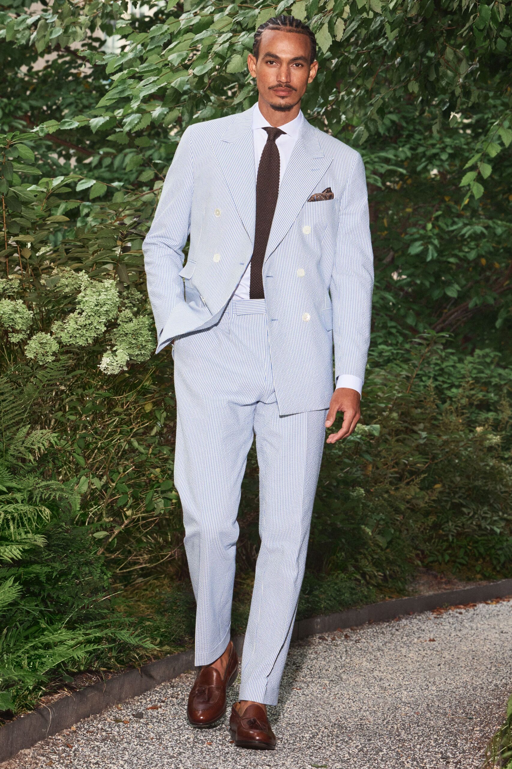 Powder-blue seersucker double-breasted jacket with broad peak lapels. White dress shirt with a crisp spread collar. Dark brown grenadine tie. Matching seersucker trousers with a cropped break above the shoe. Chestnut tassel loafers, almond-toed and glossy. Garden reception gravel crunching underfoot before the first drink.