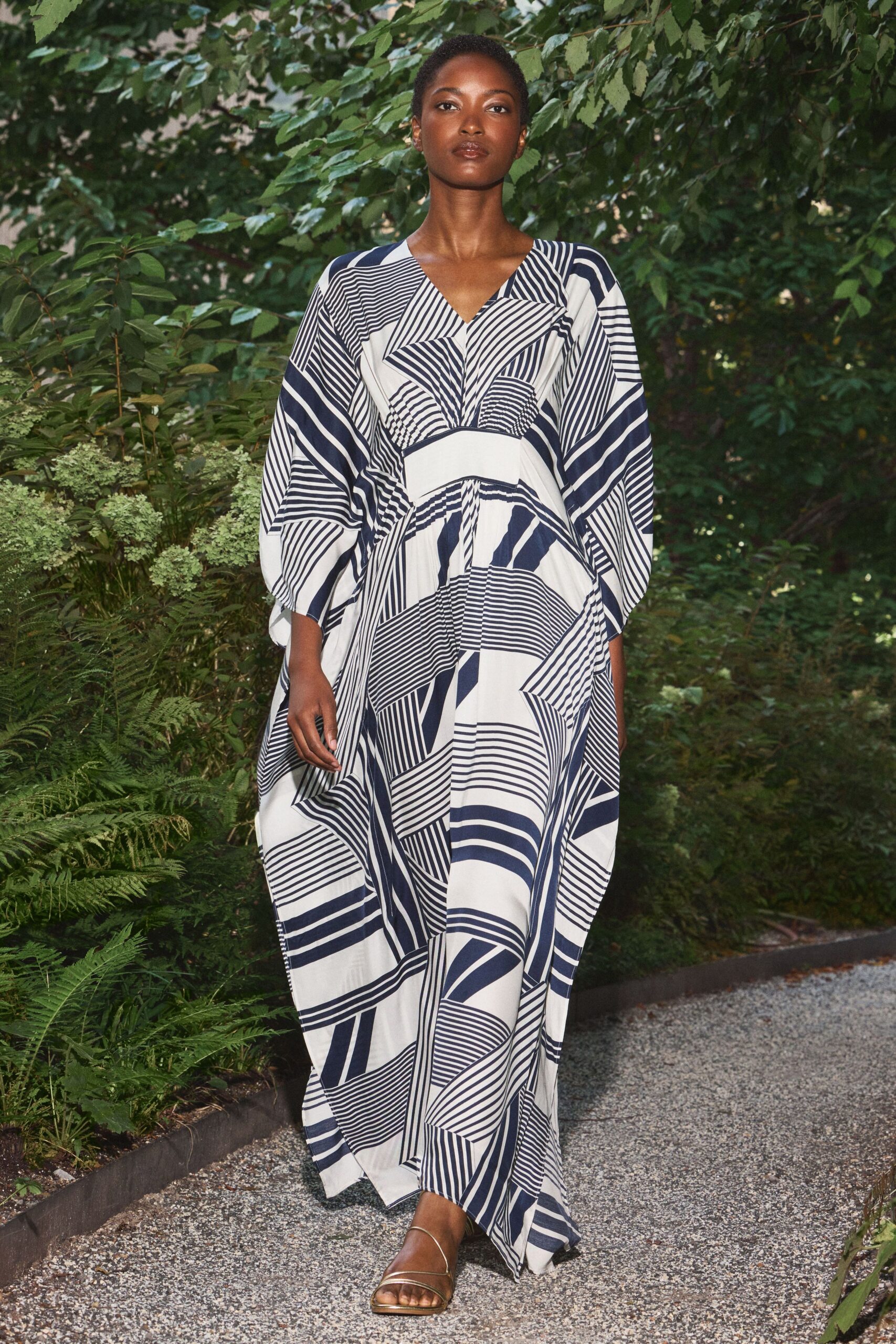 Ivory caftan dress in fluid cloth, pieced with navy striped geometric panels and dolman sleeves. Flat metallic sandals with slim straps crossing low over the toes. Walking a gravel garden path just before the air turns cool.
