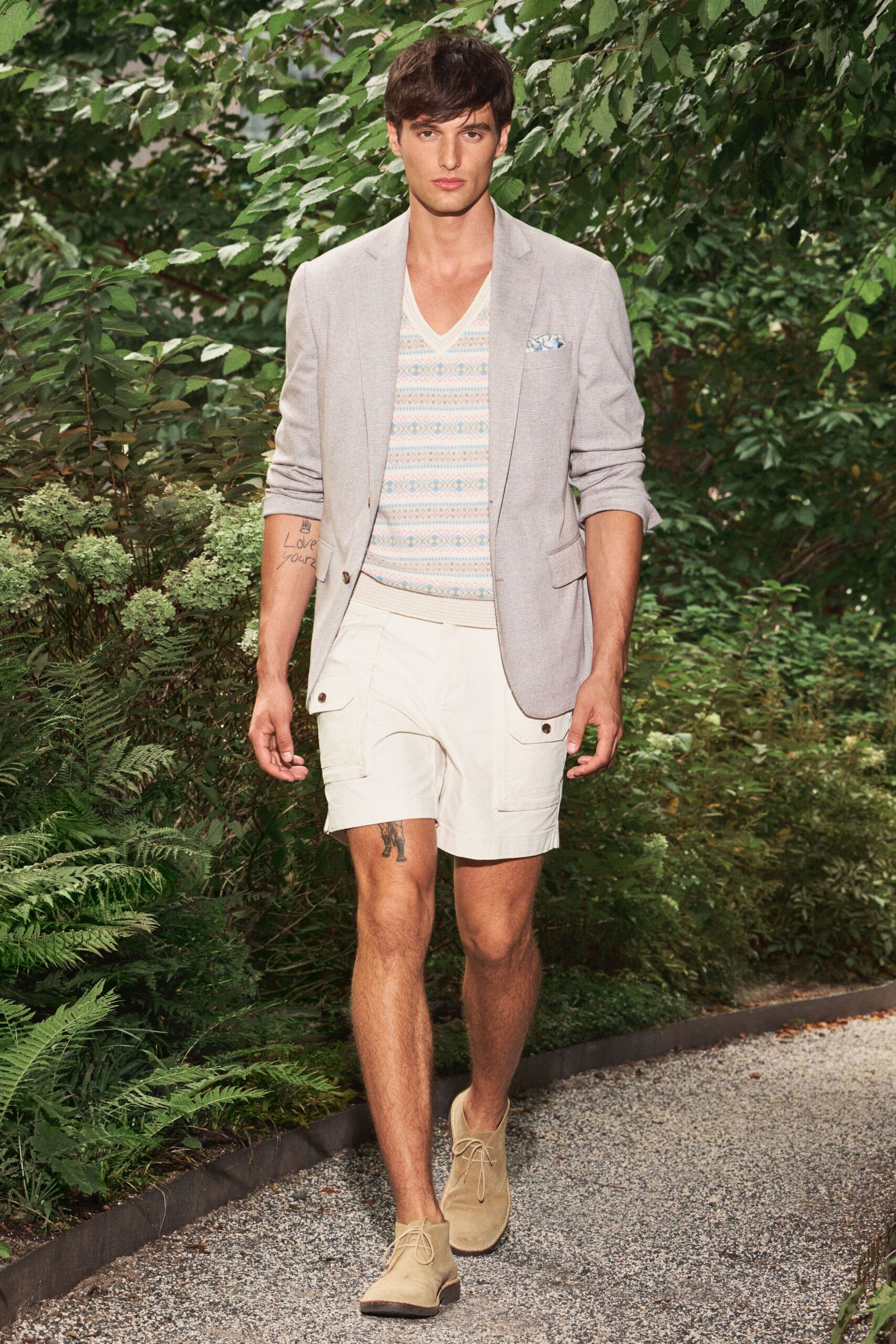 Pale heathered blazer, sleeves shoved up and a blue printed pocket square. Cream V-neck knit with narrow pastel fair-isle bands. Stone shorts with flap cargo pockets and dark snap corners. Sand suede desert boots, crepe soles dusty at the edge. Gravel garden path after brunch, still carrying the lake-house dress code.