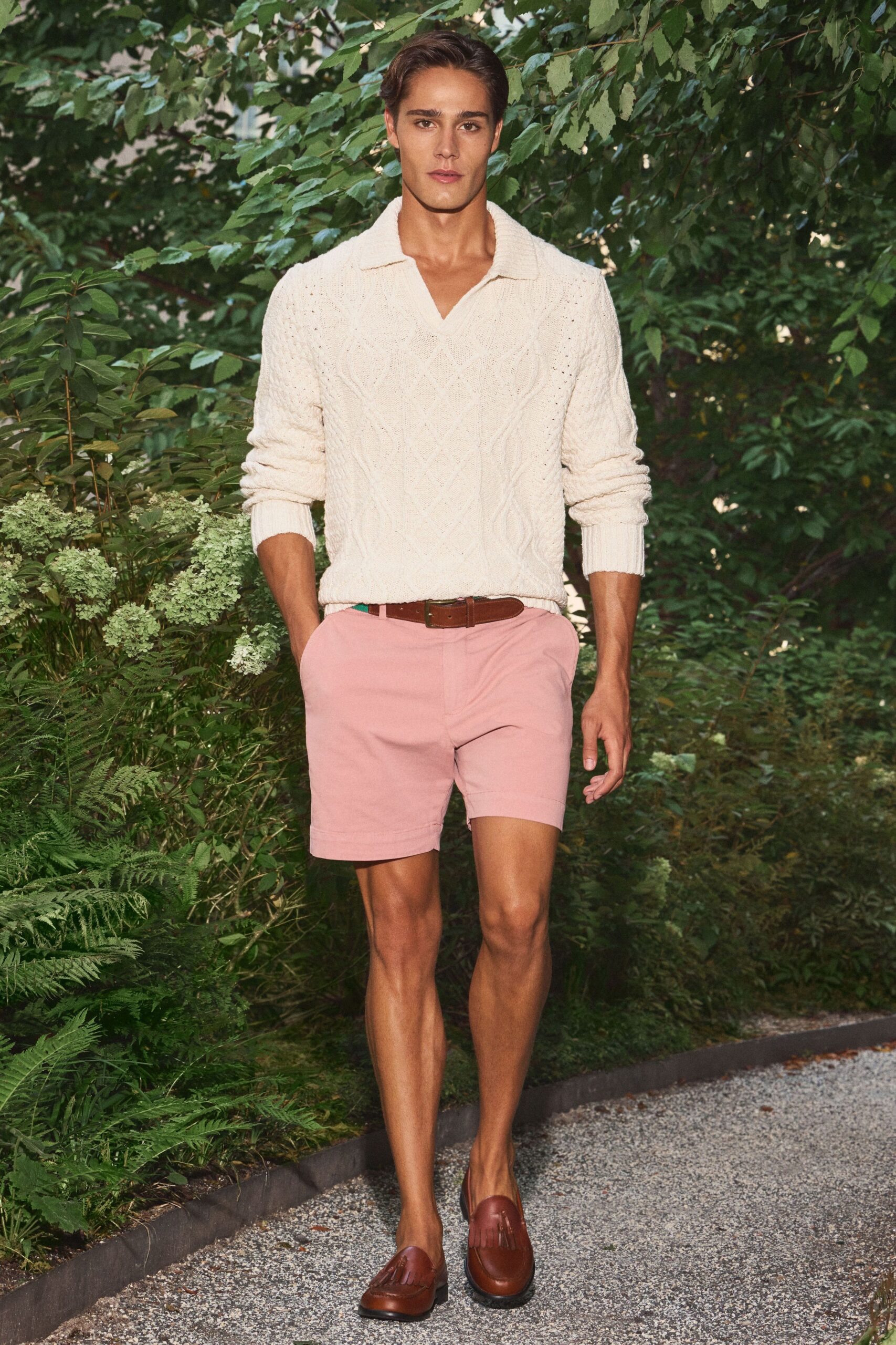 Cream cable-knit polo sweater with a deep open placket and ribbed cuffs. Shell-pink tailored shorts, cut trim through the thigh. Brown leather belt with a squared brass buckle. Chestnut leather loafers finished with a dense fringe tab. Garden path after lunch, hydrangeas gone papery in the heat.