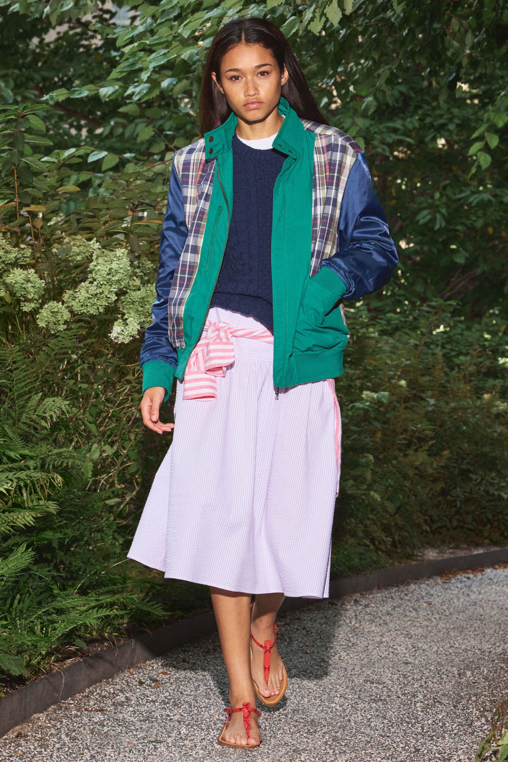 Green zip-front jacket with plaid panels and shiny cobalt sleeves. Navy cable-knit sweater, white crewneck peeking above the collar. Pale pink seersucker midi skirt with a striped shirt tied low. Flat thong sandals with thin red ankle ties. Walking the garden path after rain, still half dressed from breakfast.