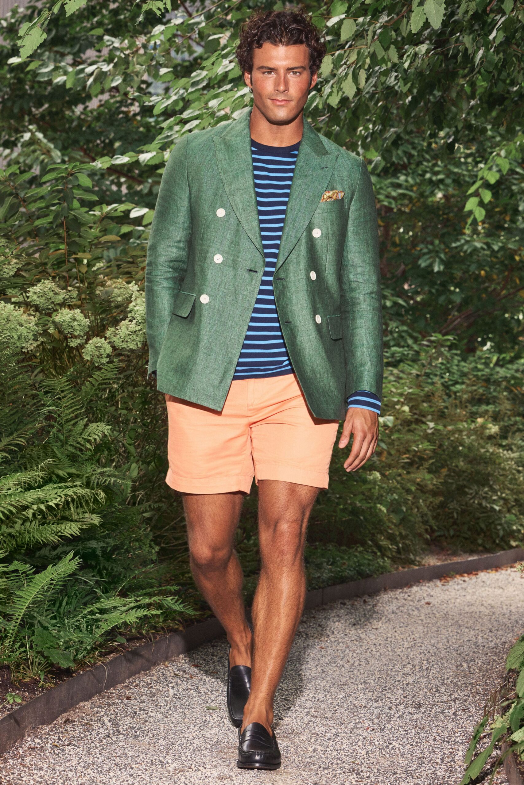 Moss linen double-breasted blazer with chalky white buttons. Navy tee banded in bright cobalt stripes. Gold paisley silk peeking from the breast pocket. Peach shorts with a clean cuffless hem. Black leather penny loafers. Walking the gravel path to lunch at a garden hotel.
