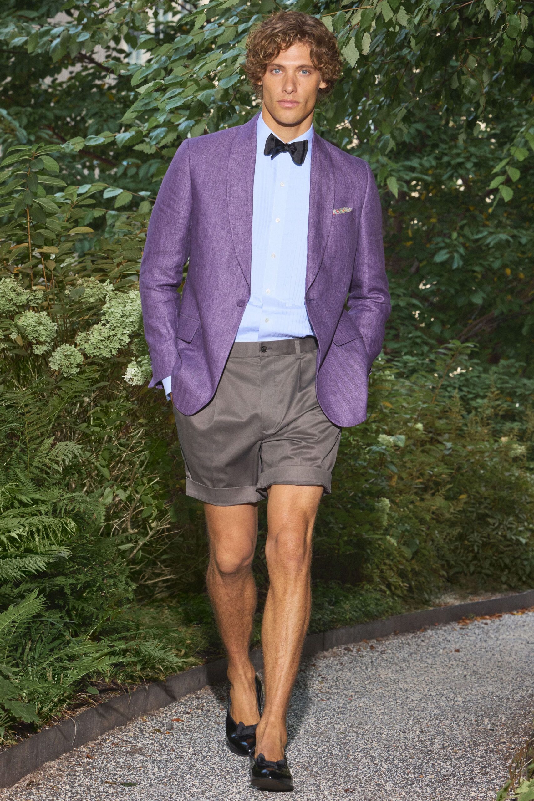 Heathered violet blazer with rounded cutaway front and flap pockets. Powder-blue bib-front shirt with fine pintucks and long cuffs. Black bow tie in matte grosgrain. Pewter tailored shorts with deep turn-up cuffs. Black patent loafers finished with neat bow trims. Garden wedding gravel, just before somebody spills champagne.