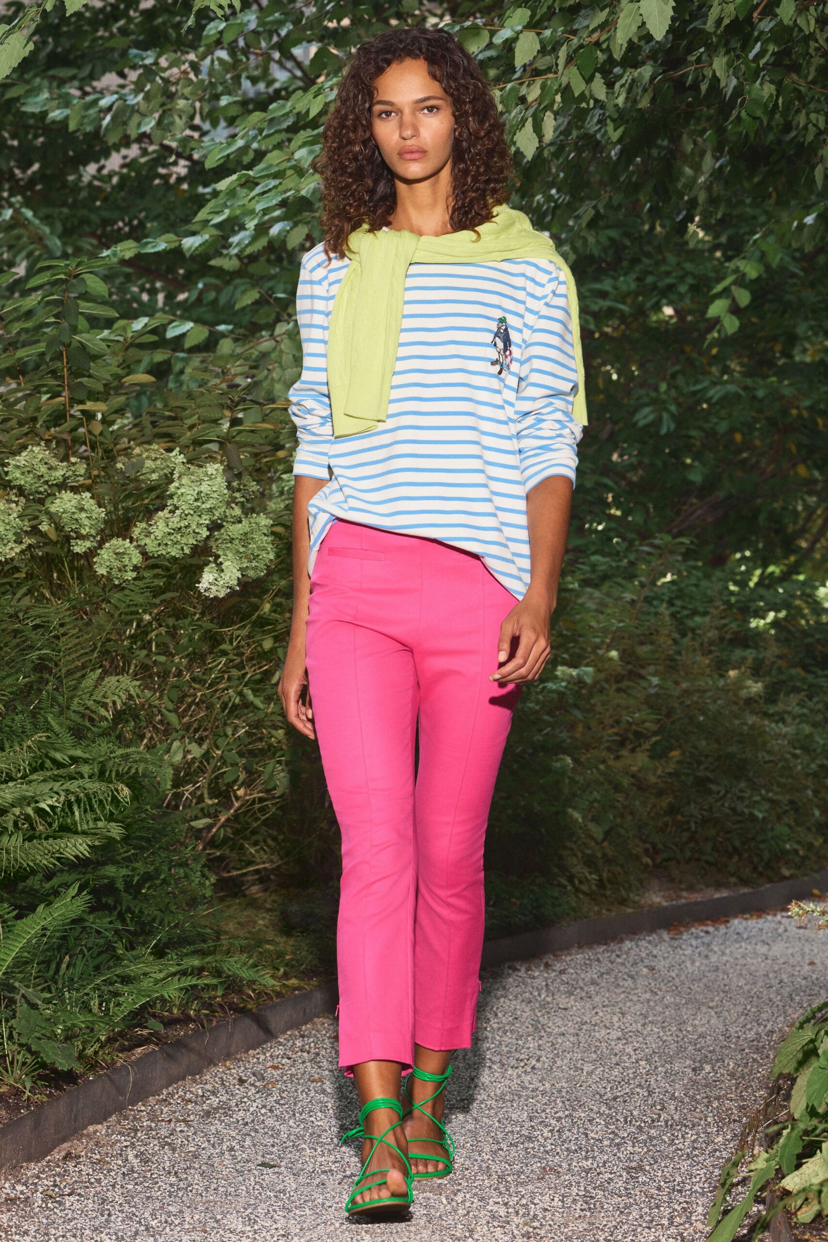 Acid-lime fine knit draped over the shoulders, sleeves knotted loose. White jersey striped sky blue, with a tiny chest figure embroidery. Hot-pink cigarette trousers with pressed seams and ankle zips. Green lace-up sandals winding thin straps over the foot. Walking the gravel path to a garden lunch that started without you.