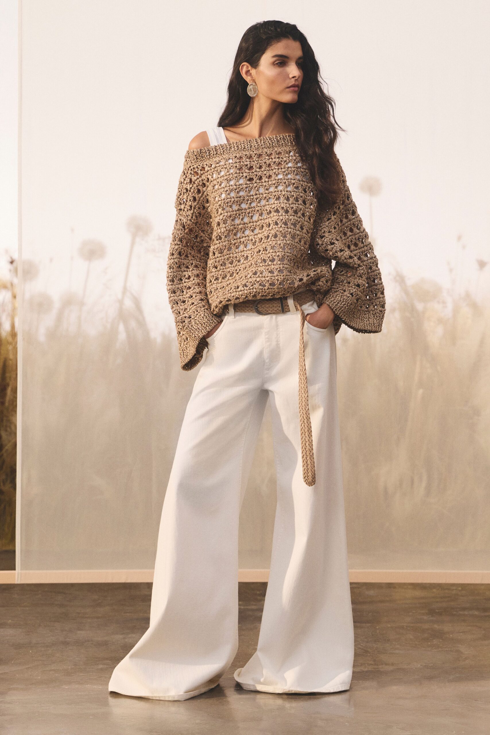 Taupe open-crochet sweater slipping off one shoulder, sleeves pooling wide. White ribbed tank strap showing at the neckline. Ivory wide-leg trousers with a heavy floor-grazing break. Braided tan belt, long end hanging past the hip. Hammered silver disc earrings. Walking through the beach house before anyone opens the shutters.