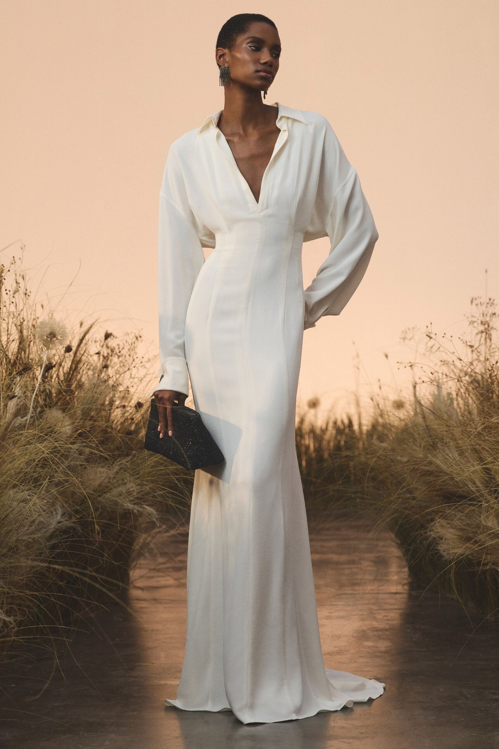 Ivory crepe gown with a plunging split collar and elongated cuffs. Black beaded box clutch, held low against the hip. Gunmetal fringe earrings brushing the jaw. Crossing a dune path at sunset before the vows start.