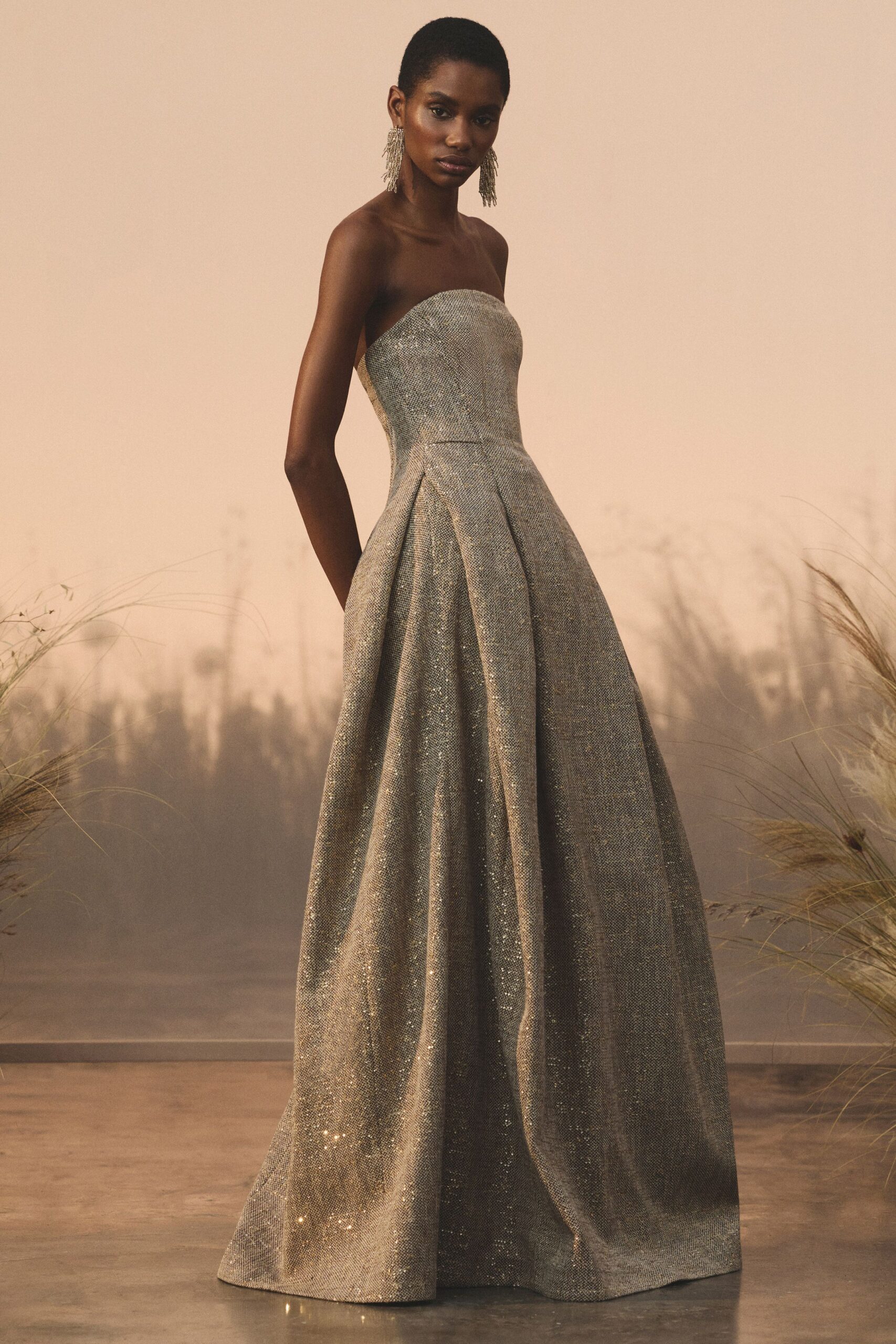Long crystal fringe earrings brushing the jaw. Strapless silver column bodice opening into a deep-pleated glitter weave skirt. Dry meadow at dusk, when the wedding band is still tuning up.