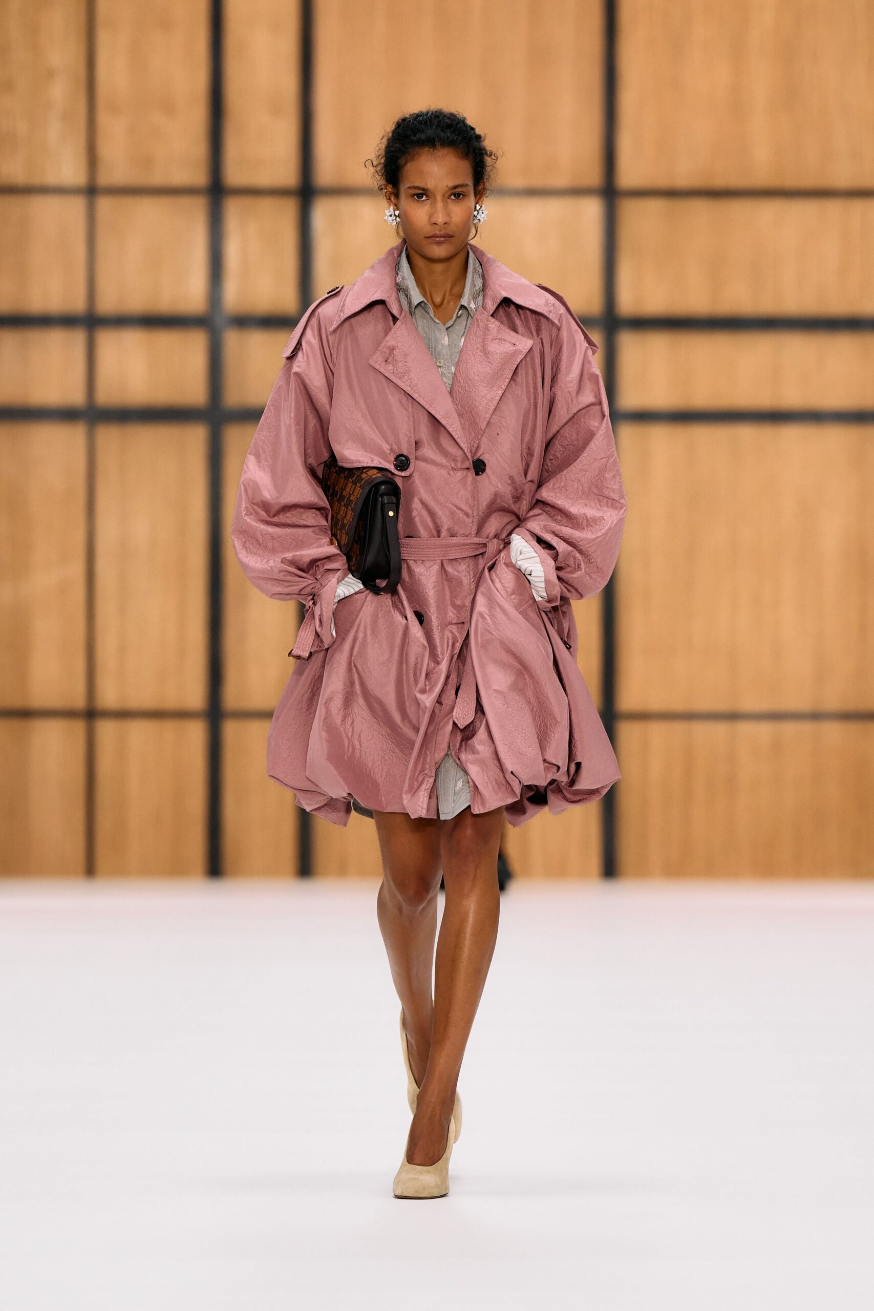 Dusty-rose crinkled trench, ballooned hem knotted with a self belt. Pale grey shirting peeking below, cuffs rolled into narrow white stripes. Brown checkerwoven flap bag tucked tight under one arm. Cream suede pumps with a softly squared toe. Crossing a hotel atrium before breakfast service starts.