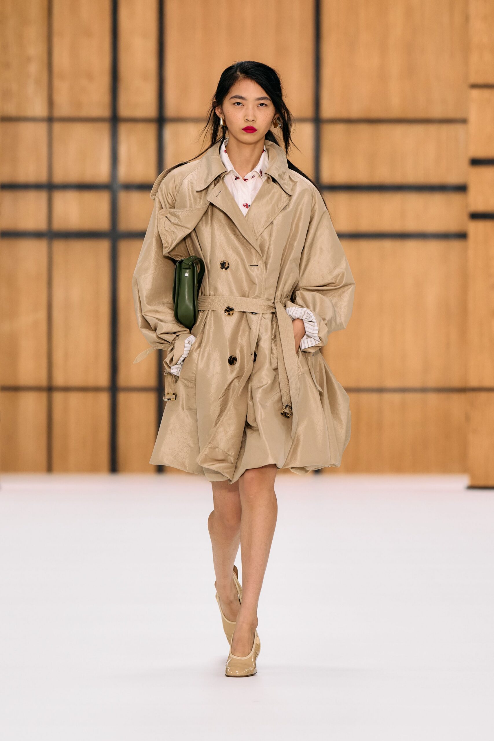 Crinkled beige trench, balloon sleeves and a loose belt with metal-capped end. White shirt scattered with tiny red floral sprigs. Olive leather clutch tucked under the arm. Cream patent pumps with a squared throat. For crossing a quiet hotel lobby before the breakfast trays are cleared.